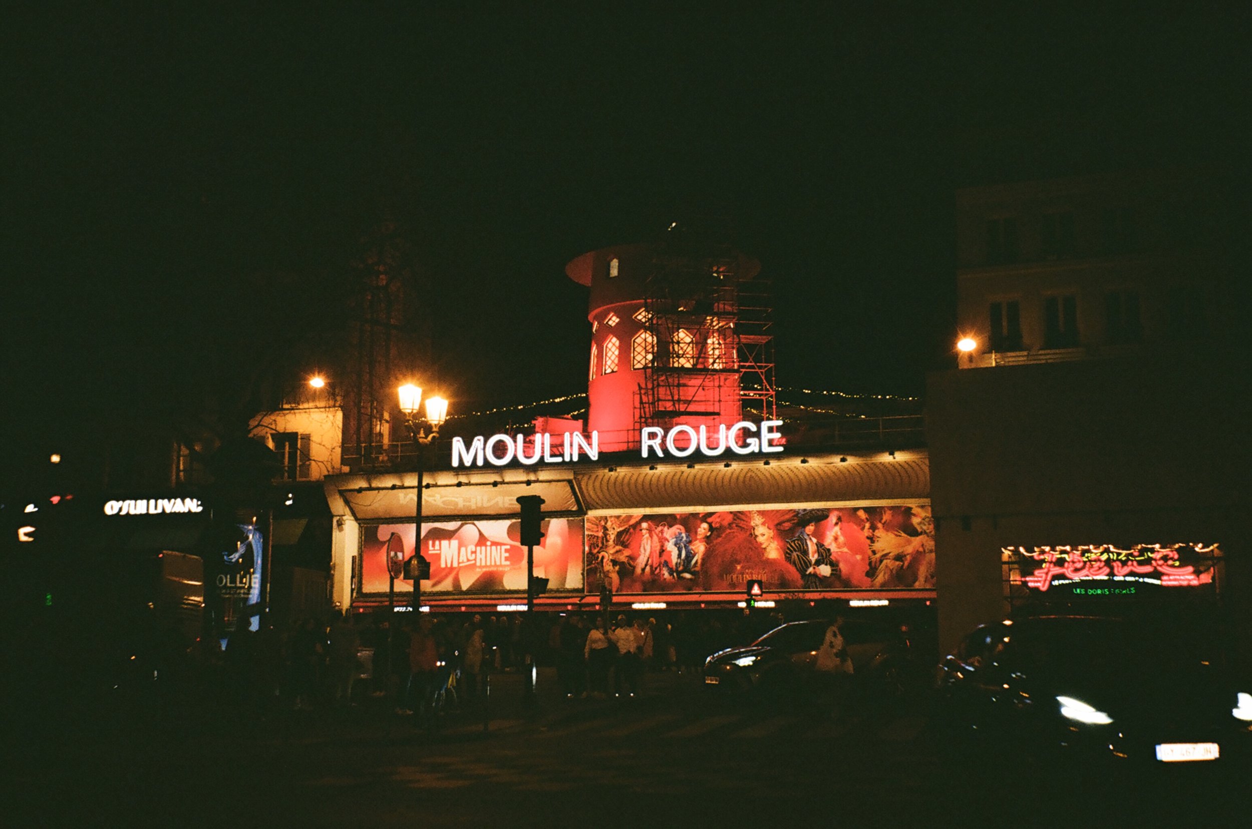 Night view of the illuminated Moulin Rouge sign with a red and black windmill structure on top, and posters advertising a show called "La Machine" below, in Paris.