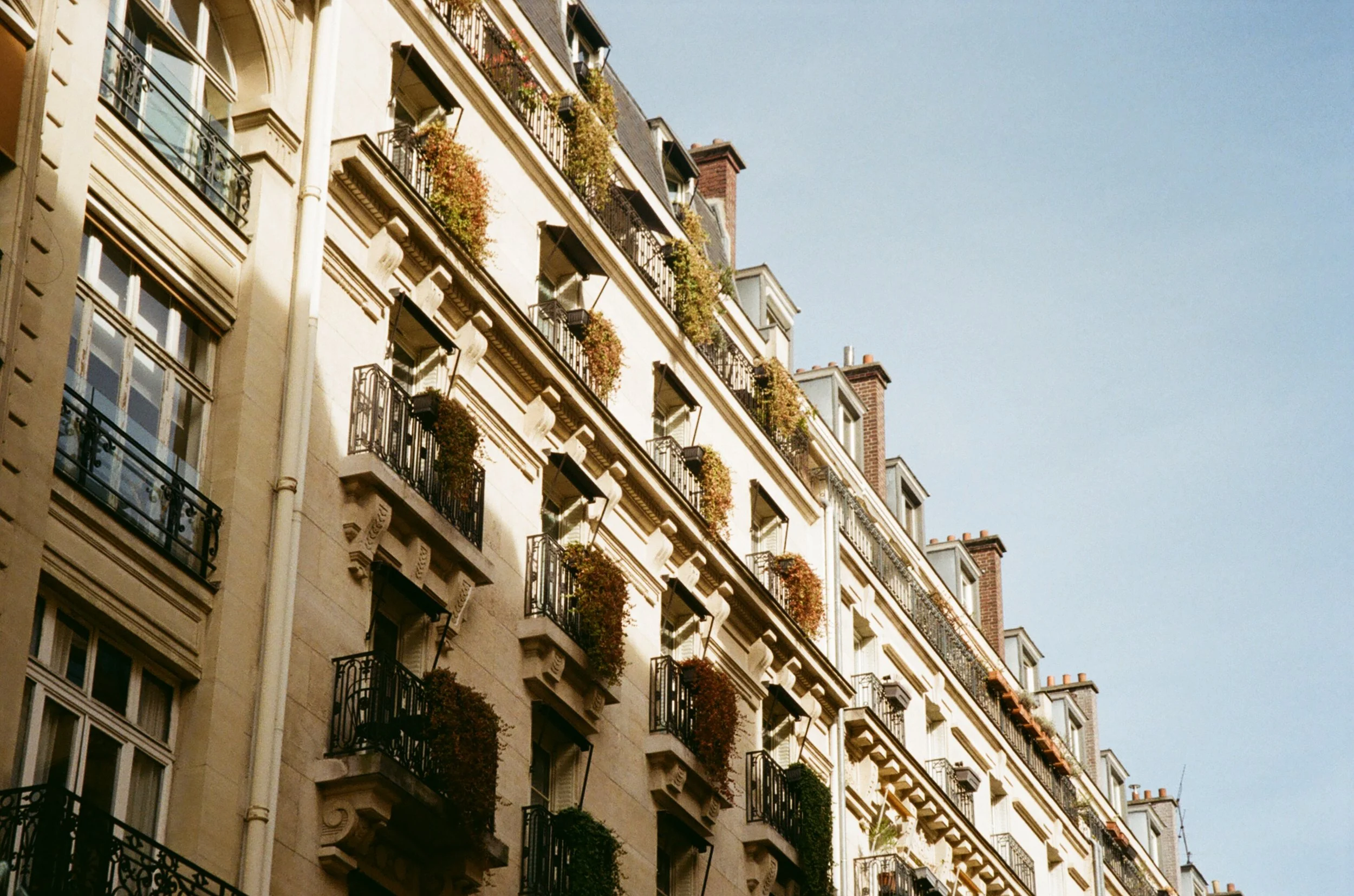 Multiple stories of a classic European-style apartment building with black wrought iron balconies and colorful flower boxes, under a clear blue sky.
