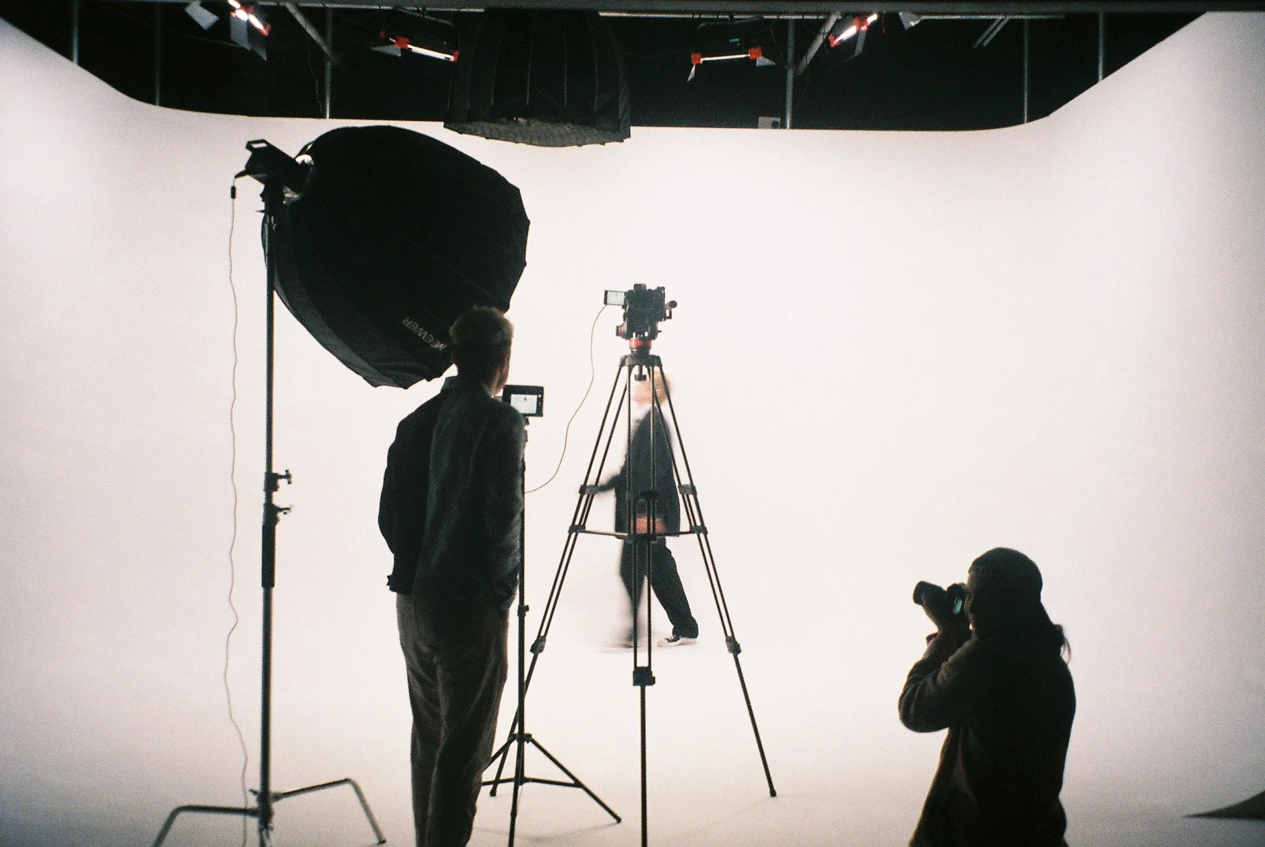 Photography studio with a white backdrop, two people setting up a camera and lighting, one person taking a photo of a model walking in the background.