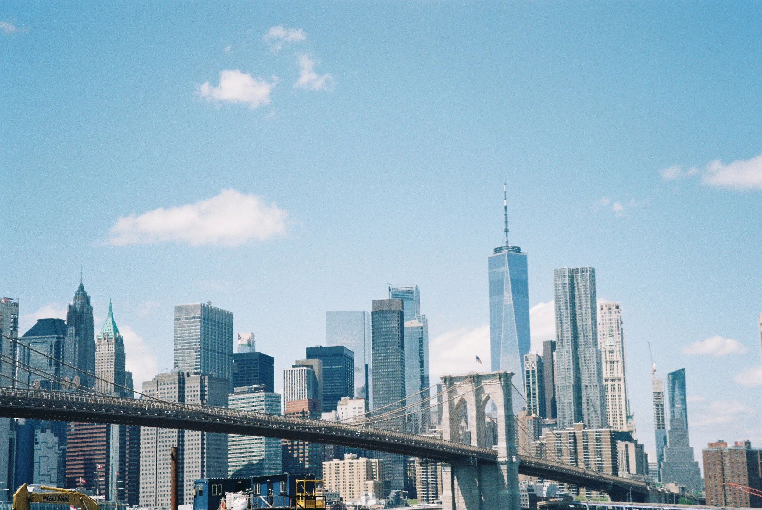 Skyline of New York City with skyscrapers, including One World Trade Center, and the Brooklyn Bridge in the foreground on a sunny day with a few clouds.