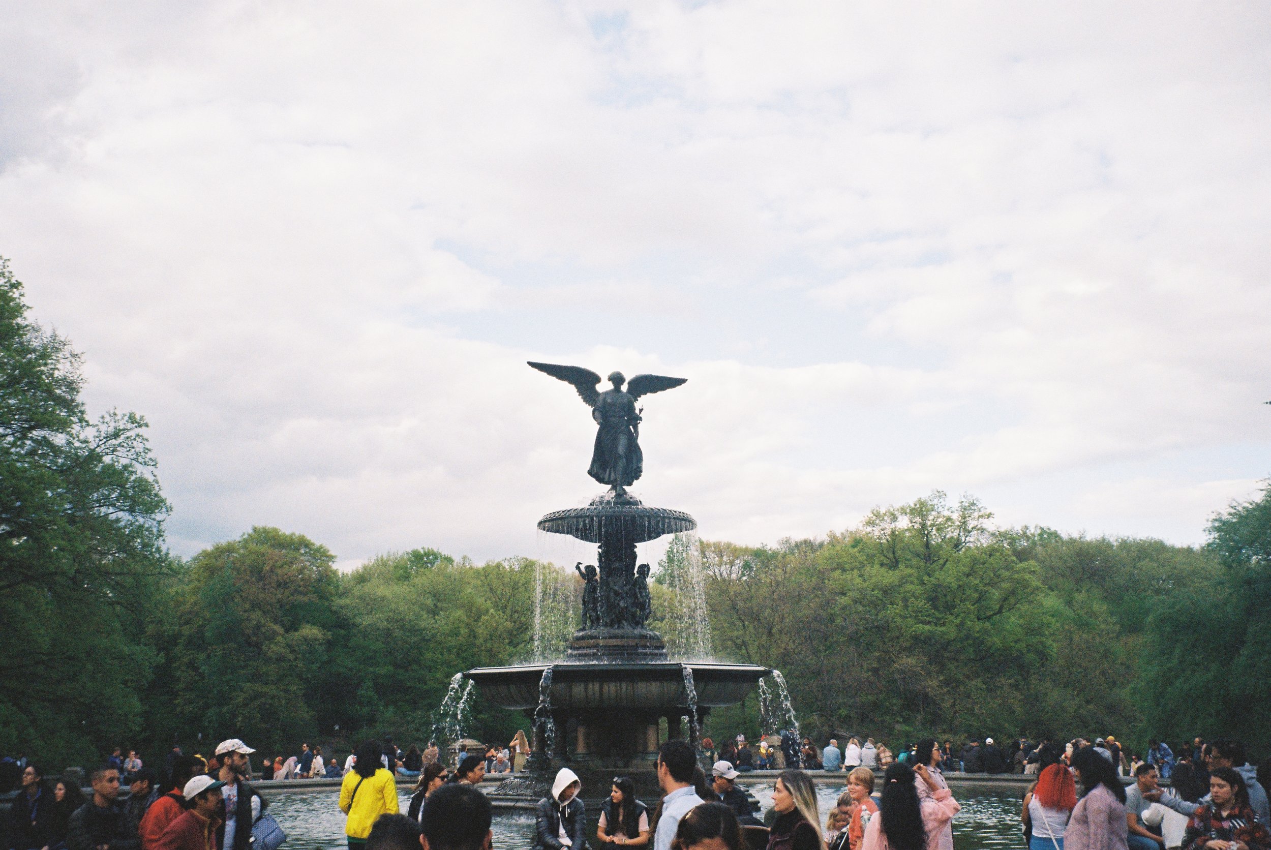 Washington Square Park fountain with a statue of a figure with wings at the top, surrounded by a crowd of people, trees, and a cloudy sky.