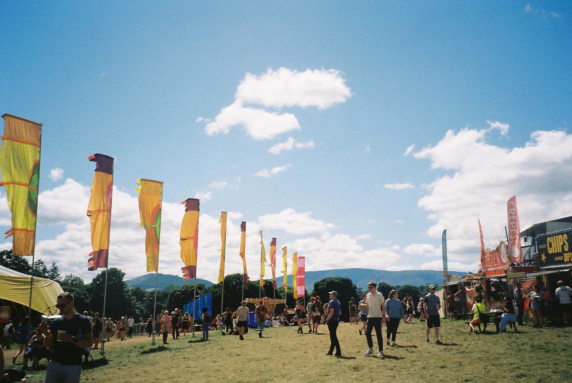 People walking and gathering on a grassy field at an outdoor festival or fair with colorful flags and food stalls, mountains and a bright blue sky with scattered clouds in the background.
