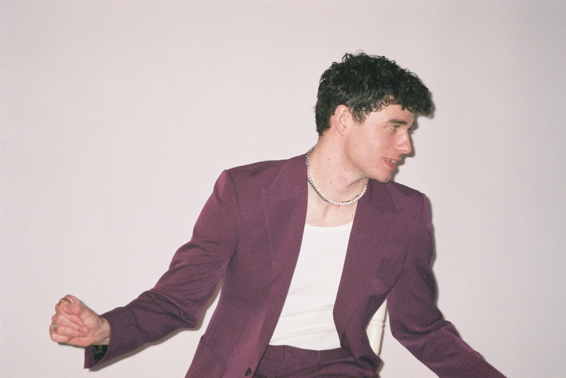 Young man with curly hair dressed in a purple suit, white shirt, wearing a pearl necklace, posing against a plain white background.
