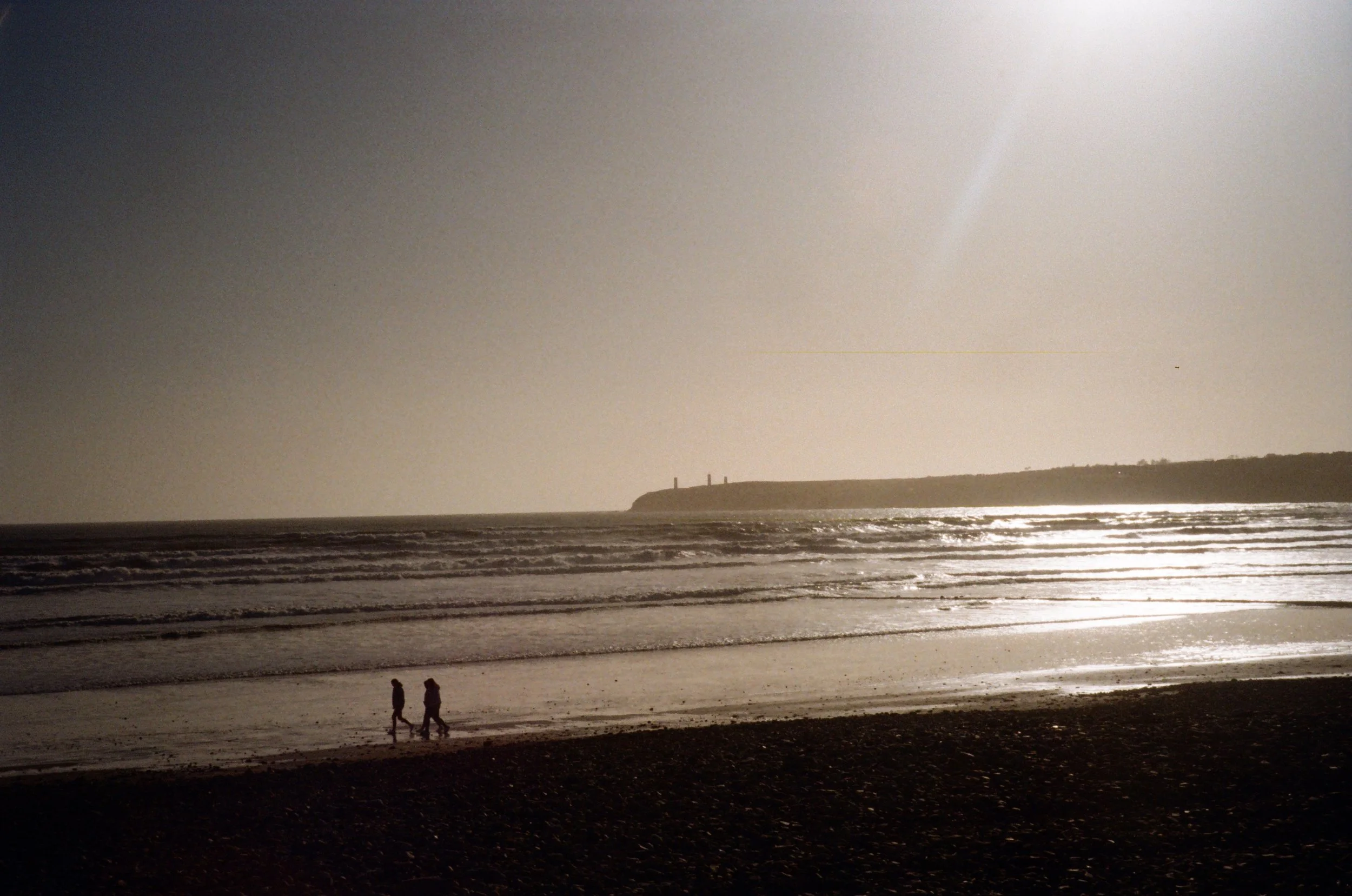Silhouettes of two people walking on the beach with waves and a distant cliff in the background under a bright sun