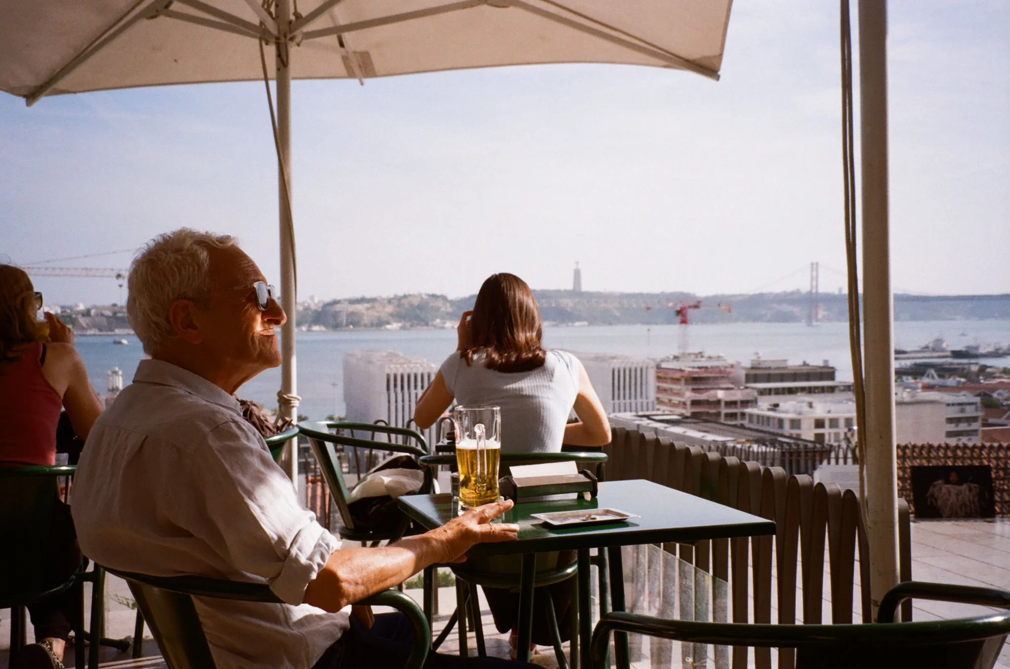 A man sitting at an outdoor table with a glass of beer, looking out over a cityscape and water with a bridge and statue in the distance, under a large white umbrella.