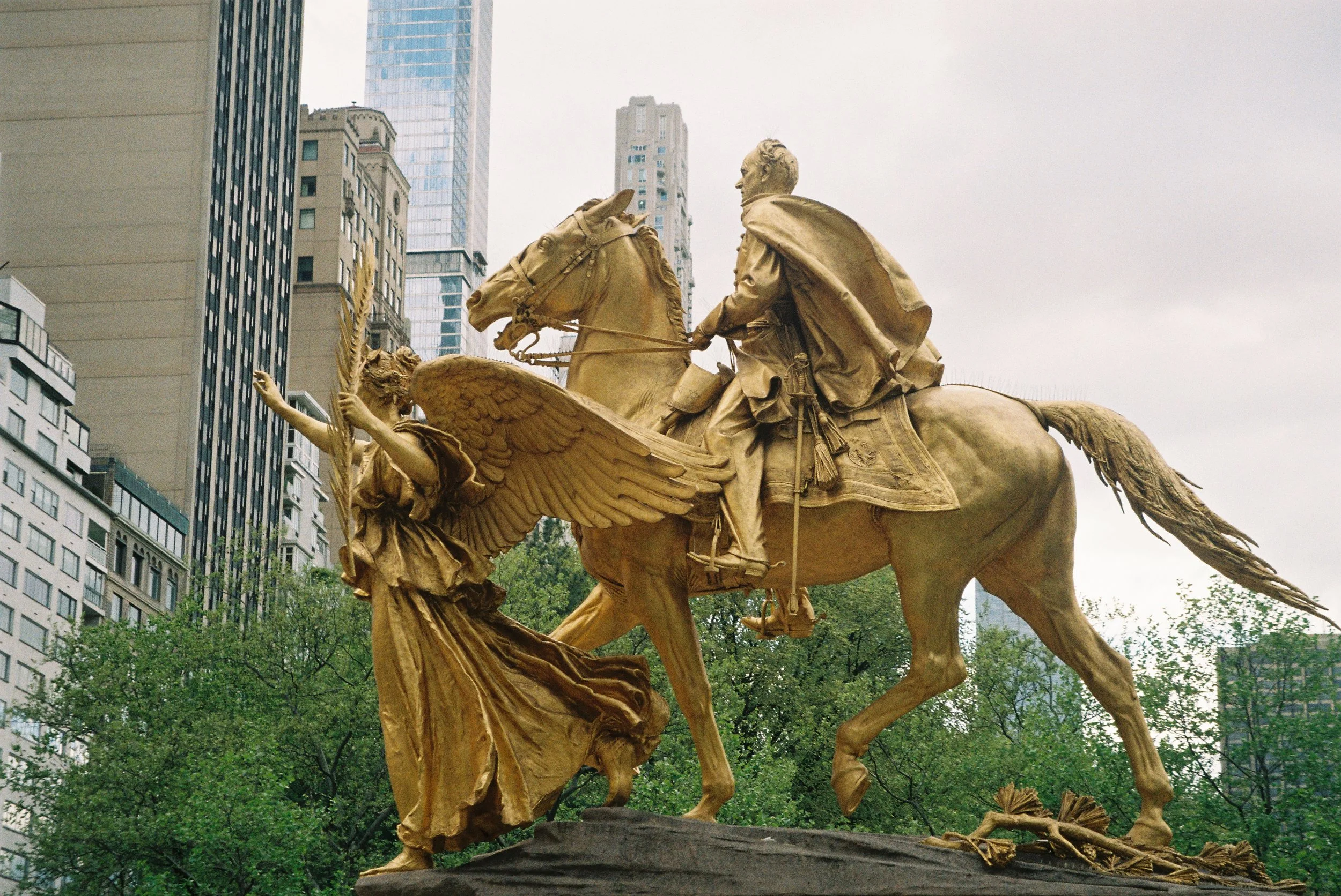 A golden statue of a man on a horse, accompanied by a winged female figure, set against a cityscape with tall buildings and greenery.