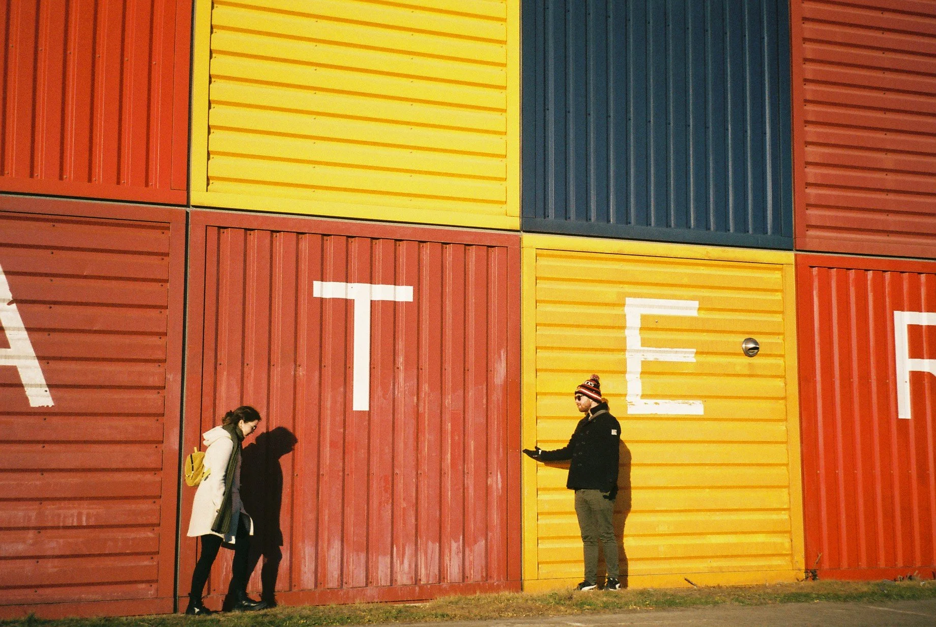 Three people standing near a colorful wall made of shipping containers in red, yellow, and blue, with large painted white letters spelling 'EAT' on the containers.