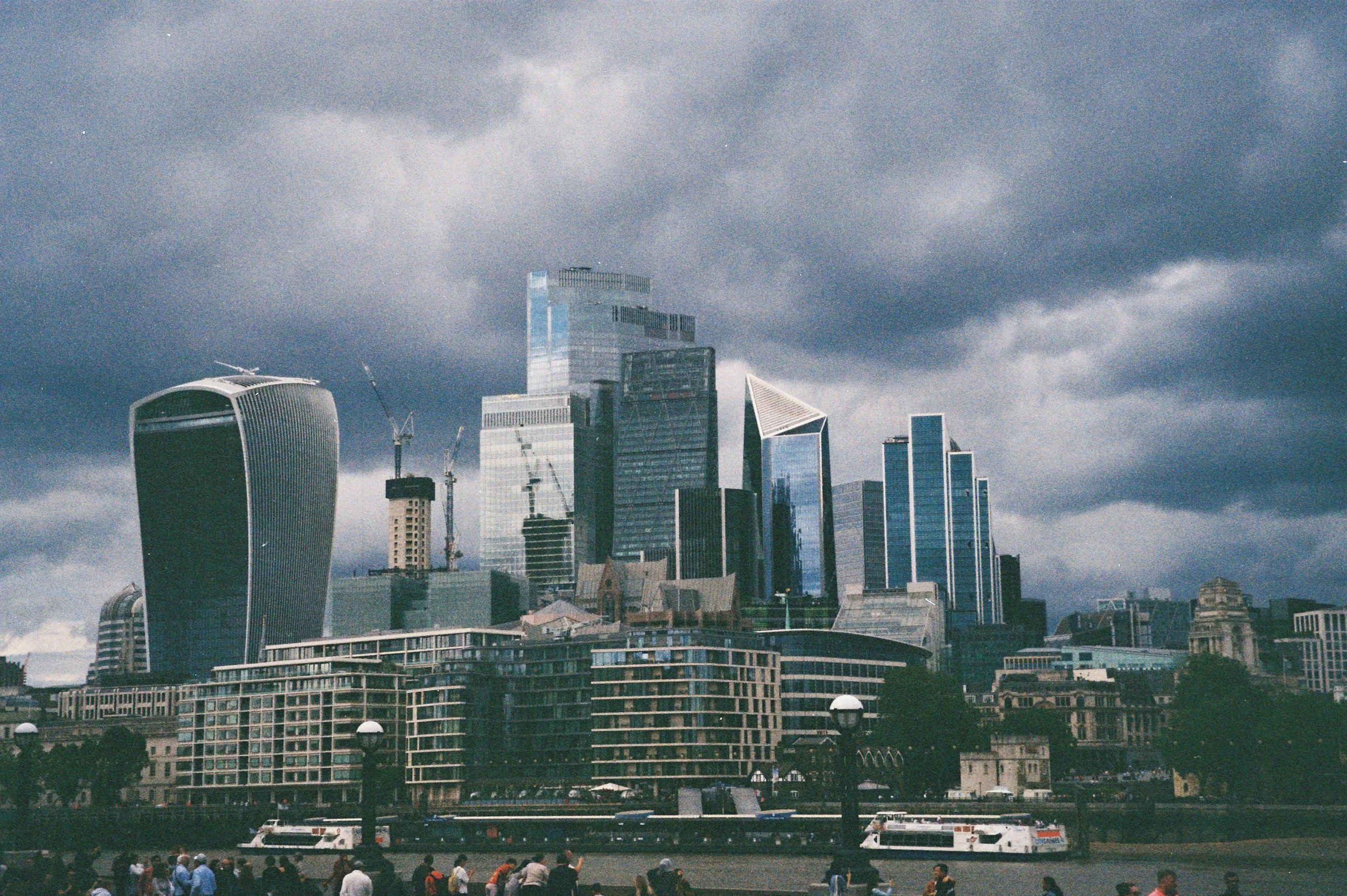 A city skyline with modern skyscrapers under dark stormy clouds, viewed from a riverbank with people and boats in the foreground.