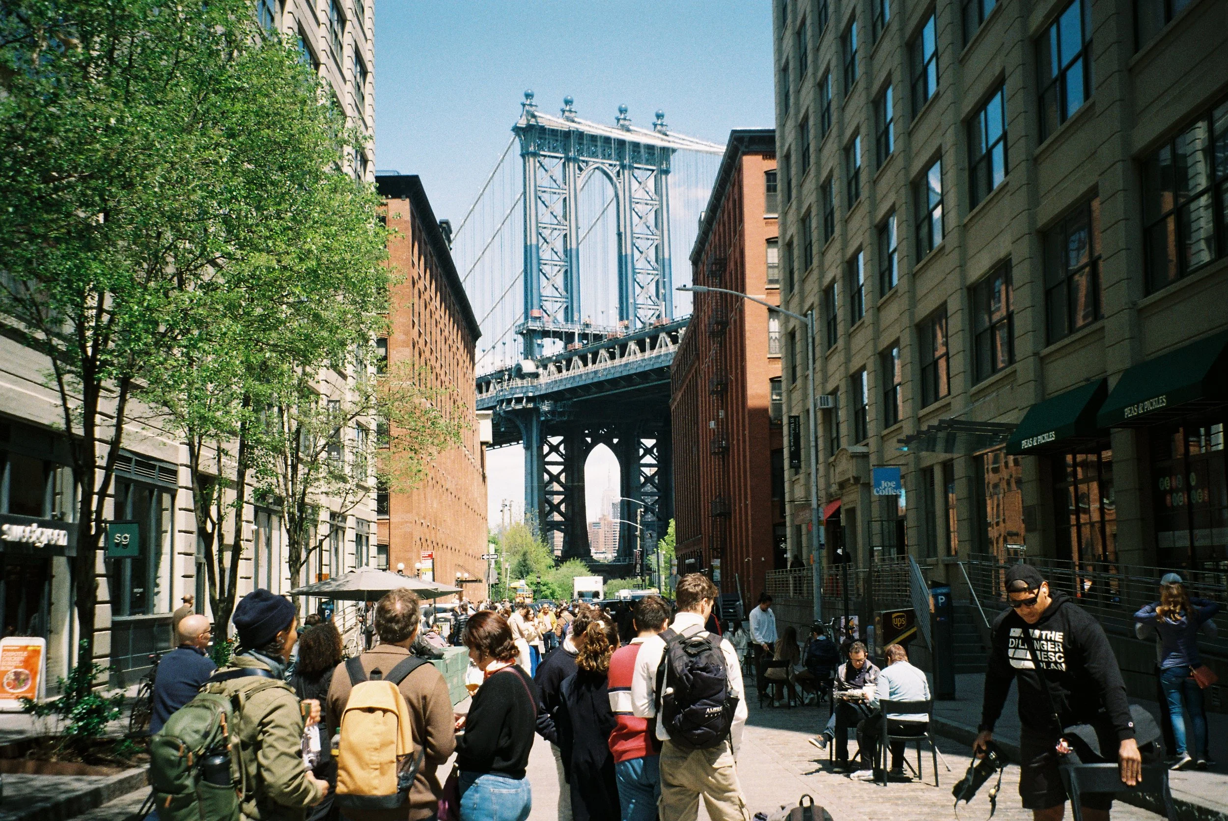 People walking and sitting at an outdoor area, with the Brooklyn Bridge and Manhattan skyline in the background.