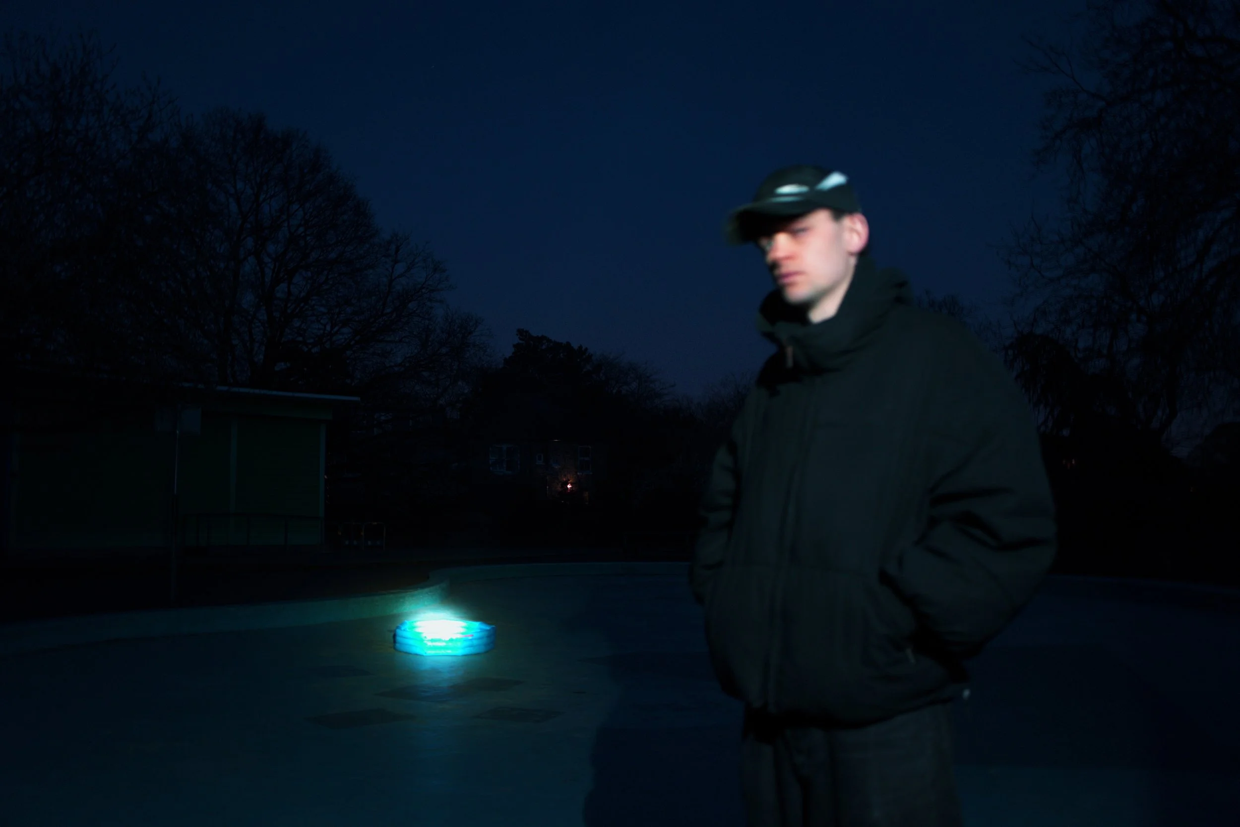 A young man in a black jacket and cap standing outdoors at night, with a glowing circular light on the ground behind him, and leafless trees in the background.