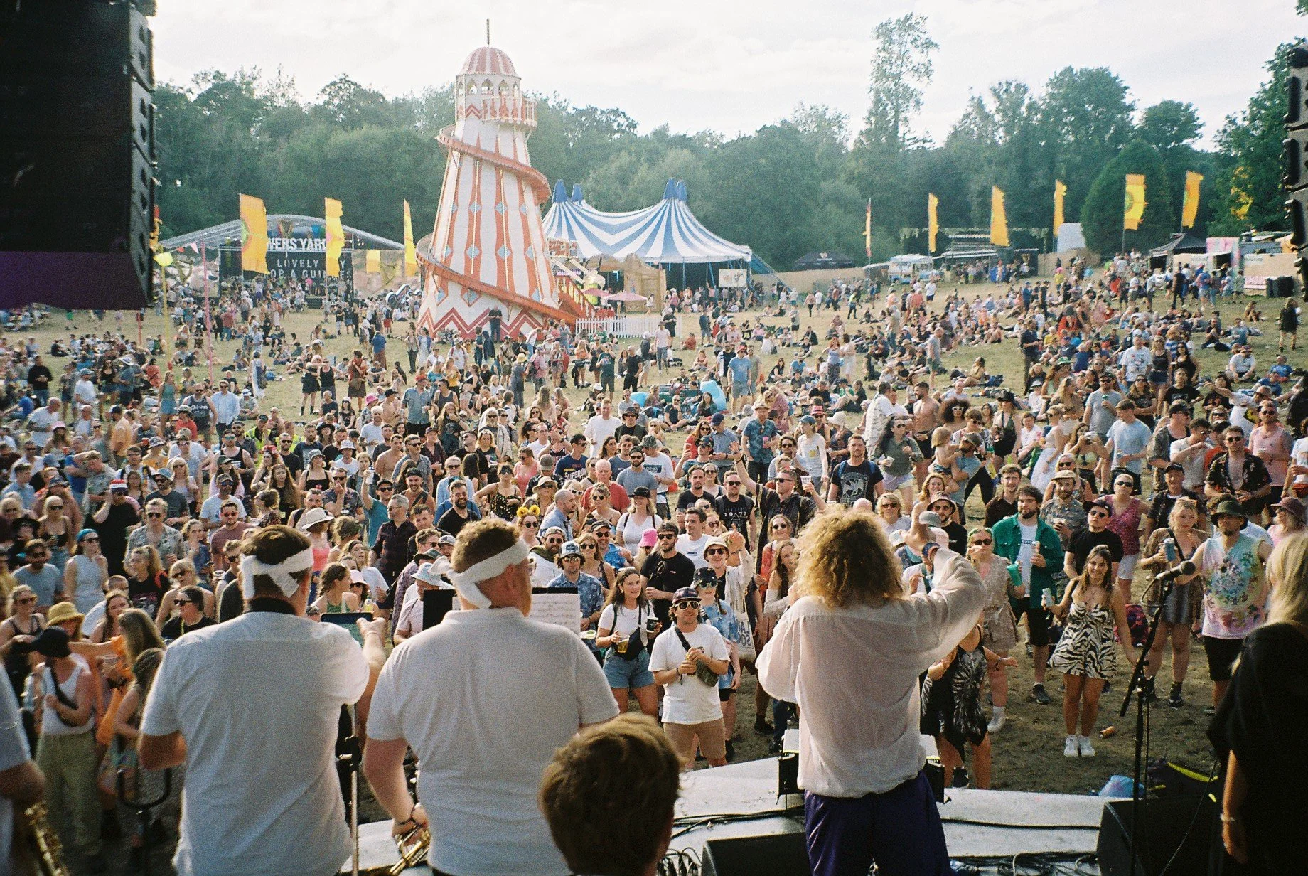 Crowd of people at a festival outdoor stage in the daytime, with various rides and tents in the background.