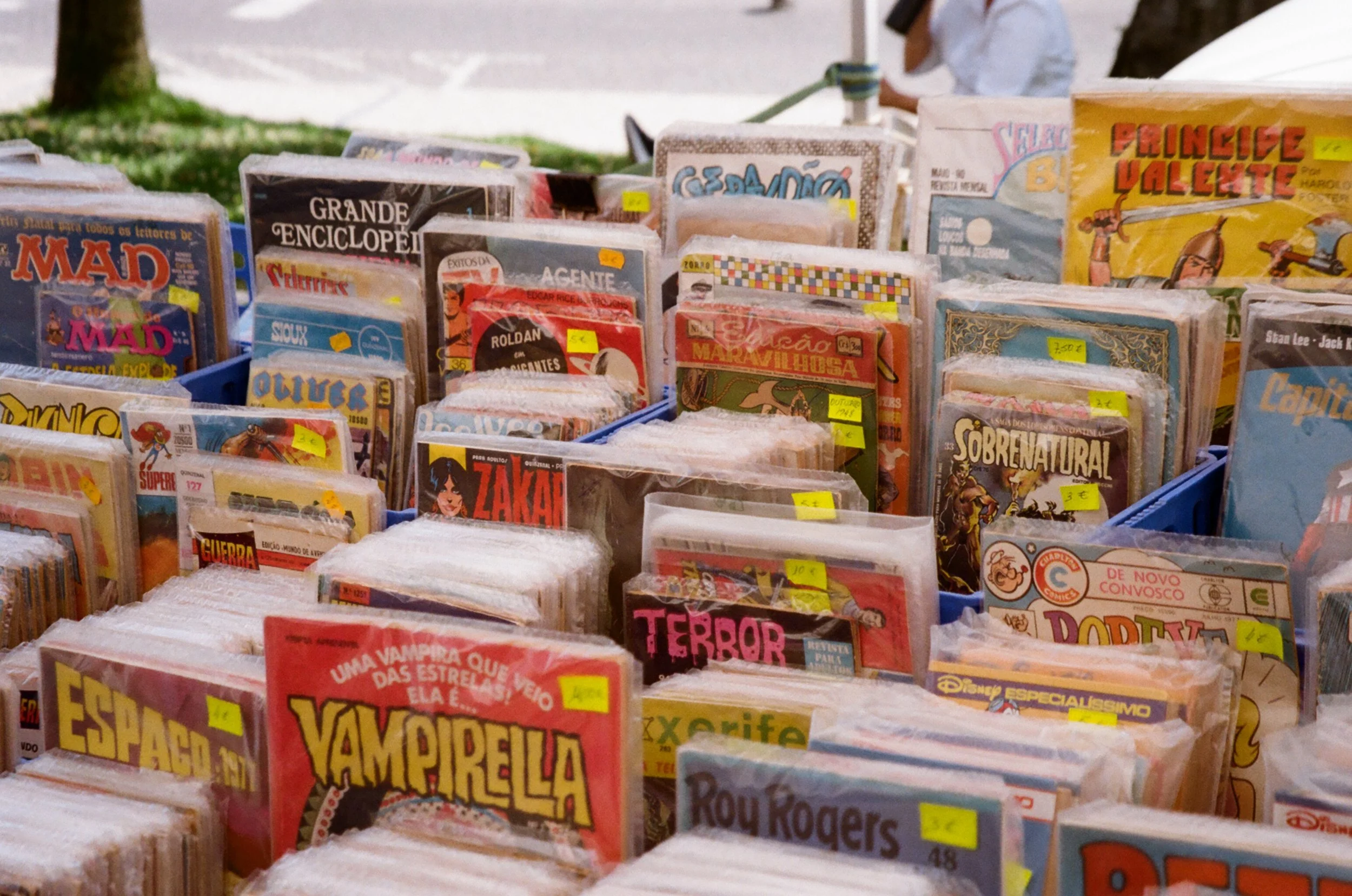 A display of comic books arranged in plastic sleeves at an outdoor market stall, with some books featuring titles like 'Vampirella,' 'Terror,' 'Super,' and 'Zara,' and yellow price stickers on each.