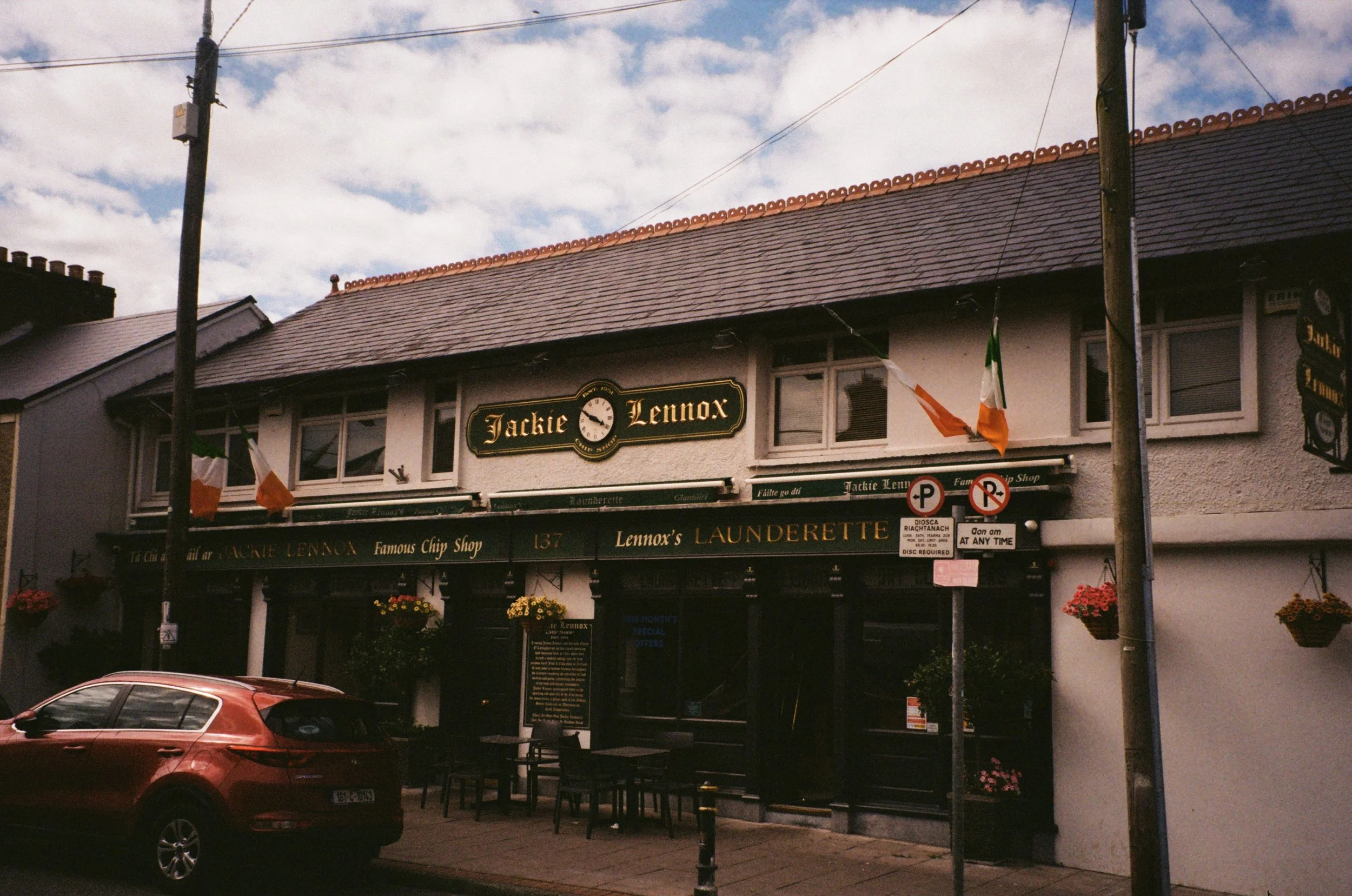 A building with a sign that reads 'Jackie Lennox' and 'Lennox's Launderette.' There are two small Irish flags hanging from the building, potted flowers on the sidewalk, a parked car in front, and street signs including 'No Parking' signs.