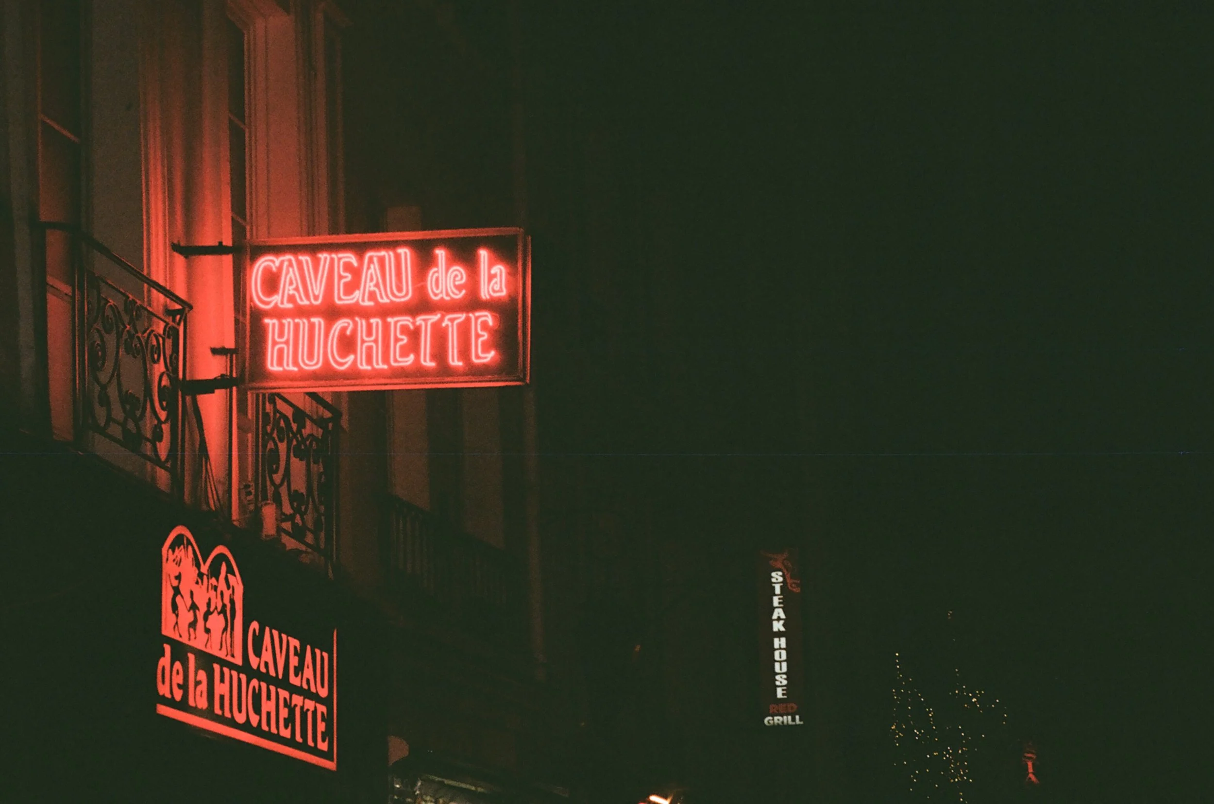 Night view of neon signs including 'CAVEAU de la HUchetTE' in red and white lights, and an adjacent sign for a steakhouse, also lit up in the dark.
