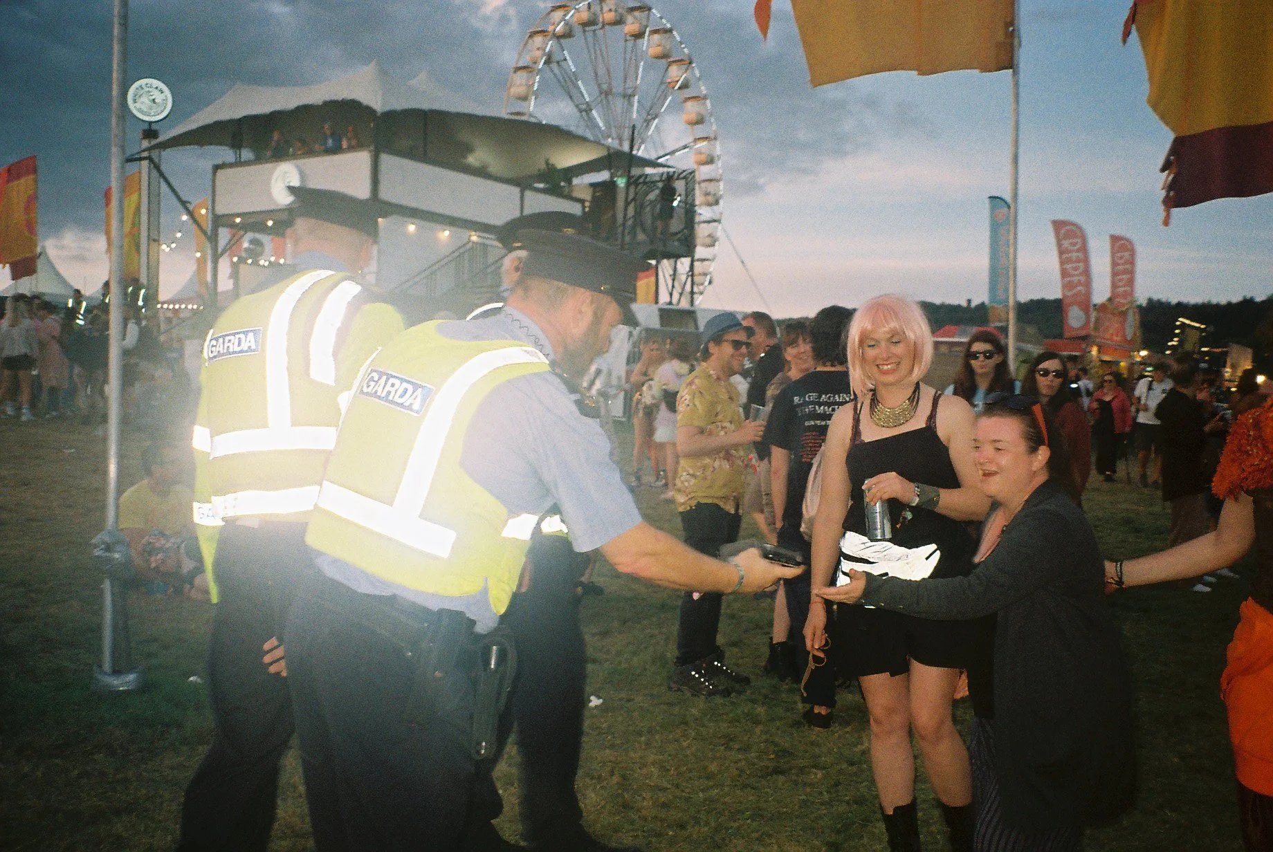 Police officers, identified by their vests labeled 'GARDA', are exchanging an item with women at a crowded outdoor festival during dusk, with a Ferris wheel in the background.