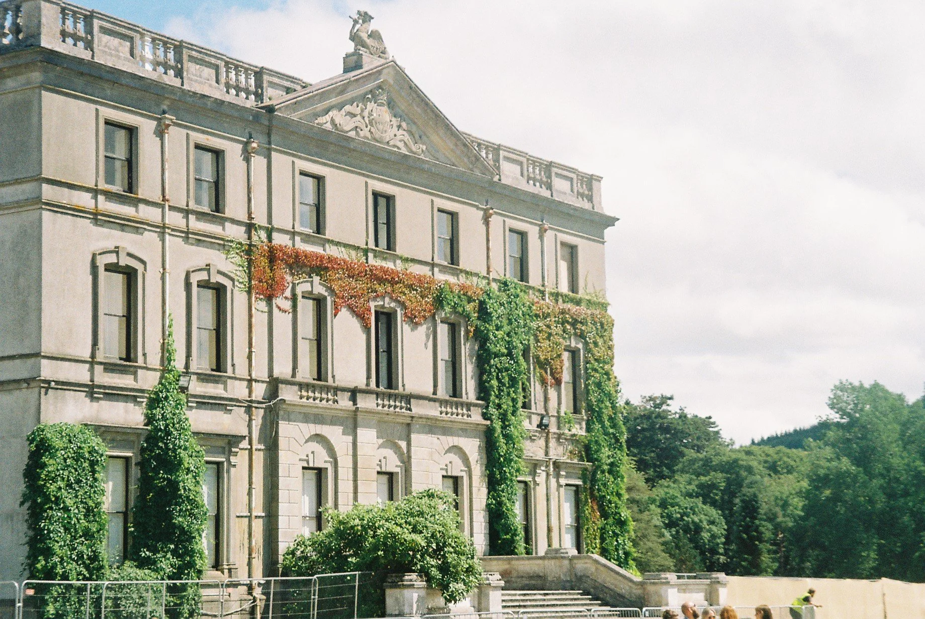 Historic multi-story building with ornate architectural details, covered partially with climbing green and red foliage, and a staircase leading up to the entrance, surrounded by trees and a cloudy sky.