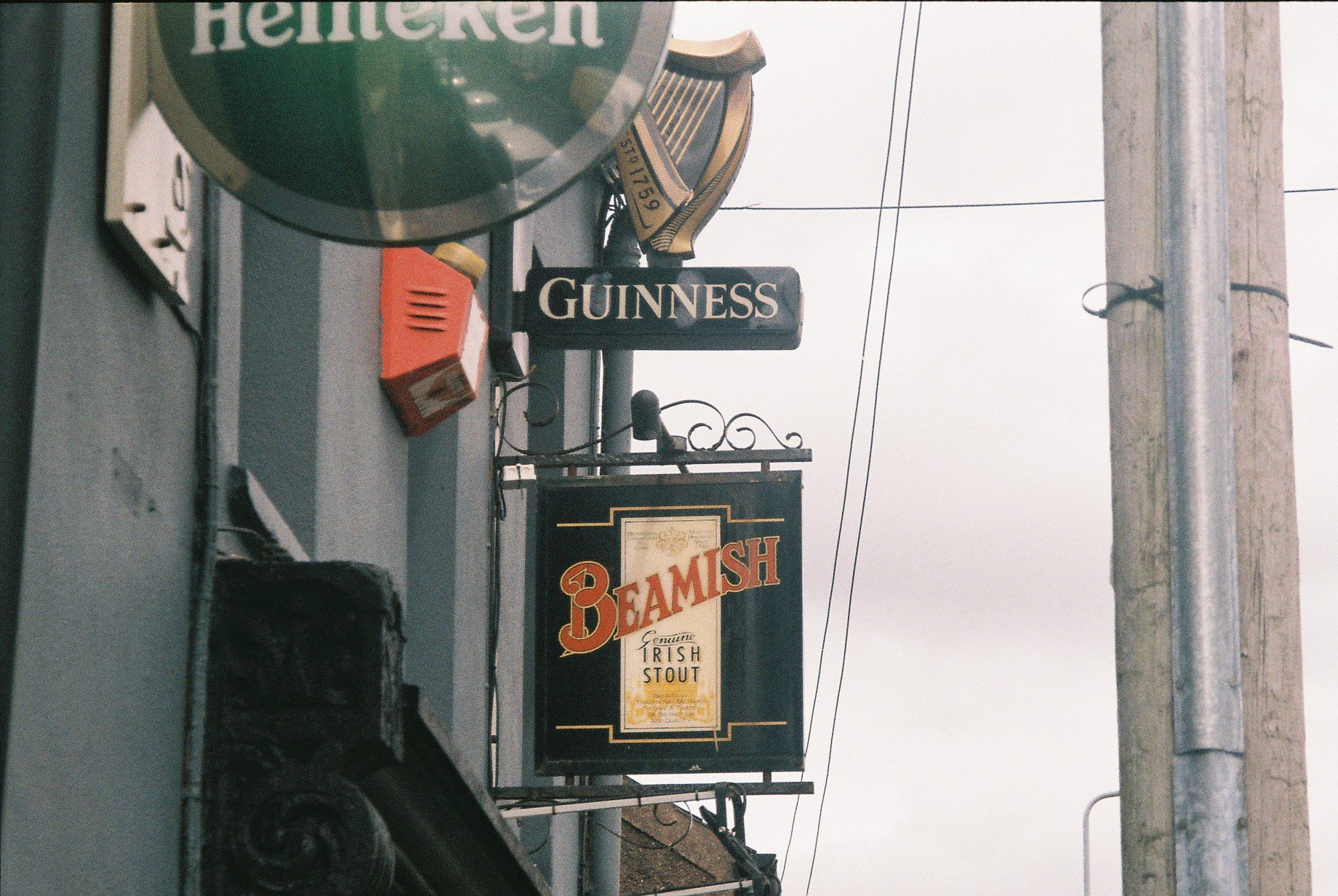 Signs on a building advertising Guinness and Beamish Irish stout beers, with a Heineken sign at the top and a utility pole with power lines against a cloudy sky.