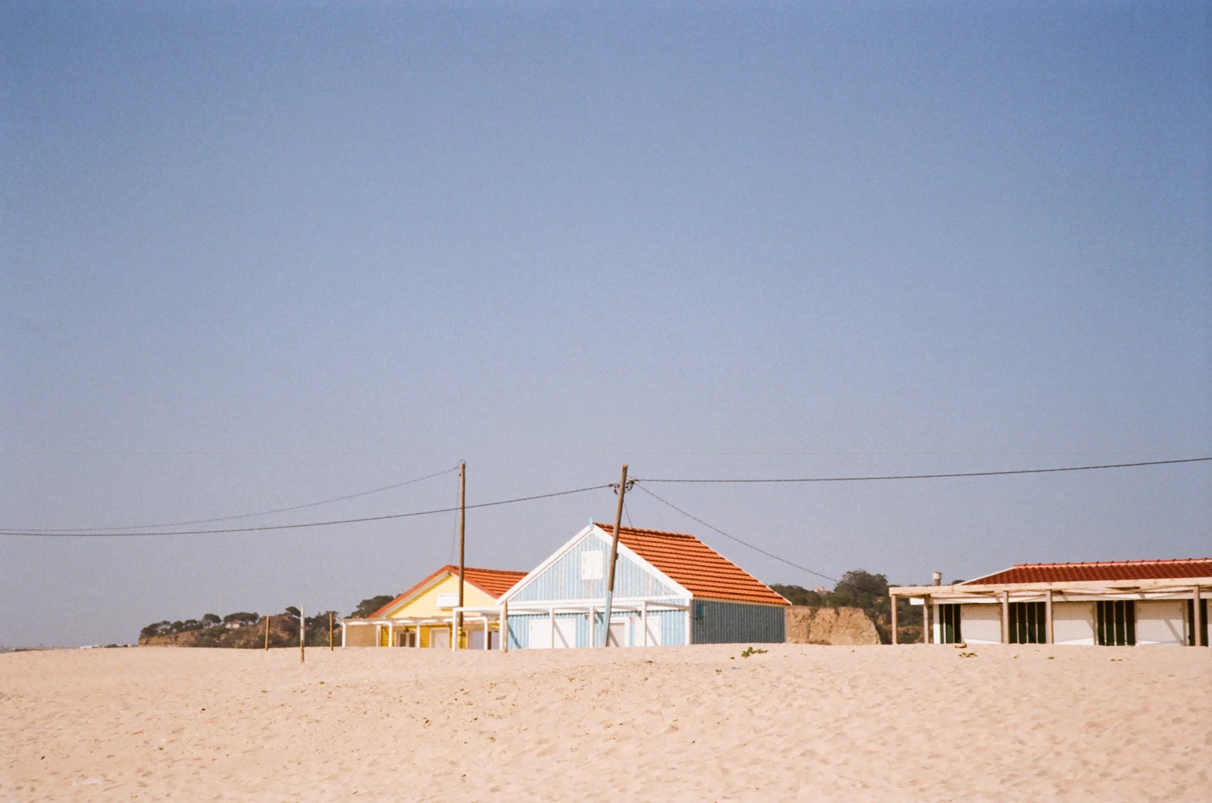 Beach with three small houses painted in pastel colors and orange-tiled roofs, sandy foreground, clear blue sky, and utility poles with wires in the distance.