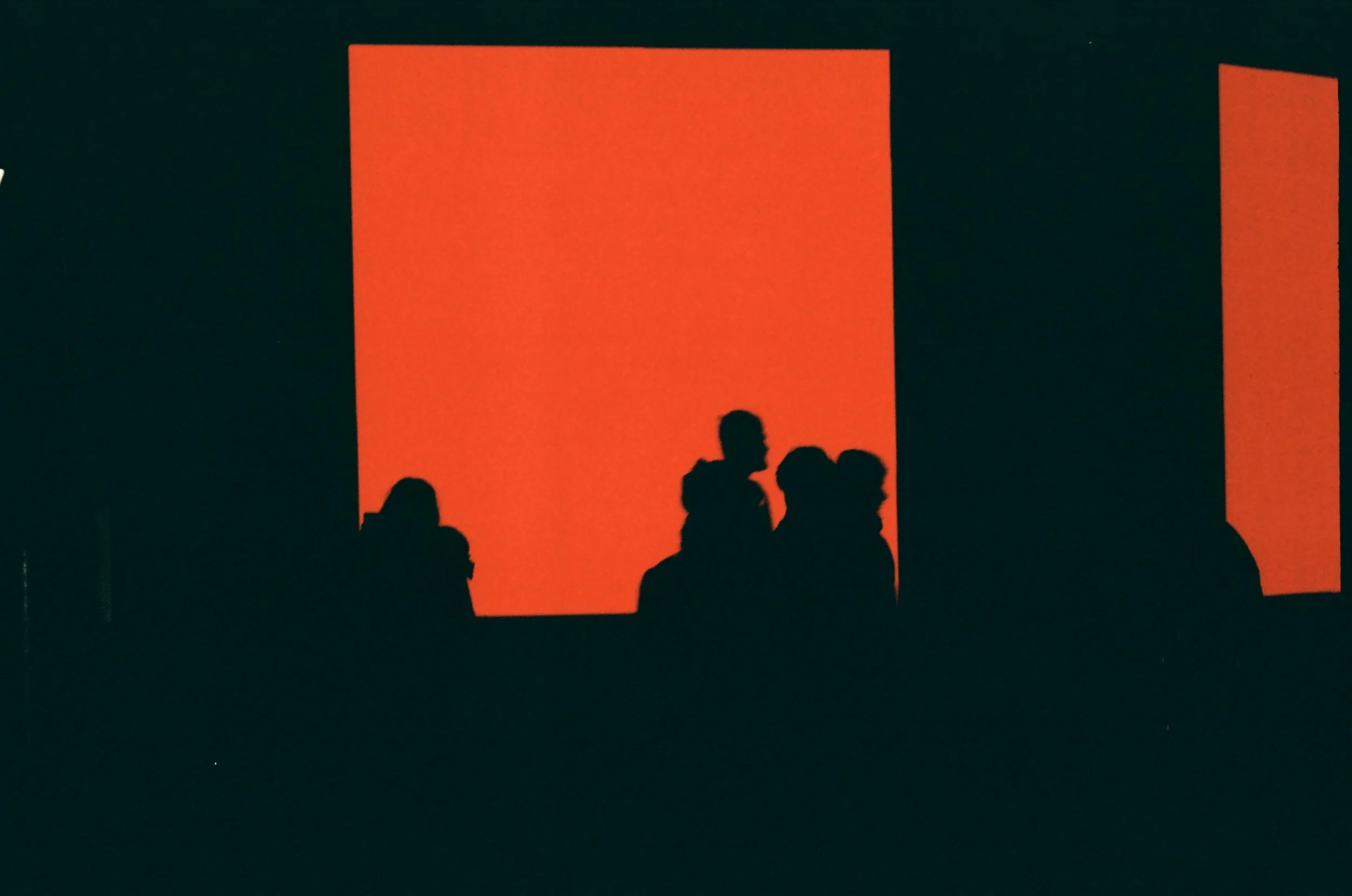 Silhouettes of people standing in front of large orange rectangular panels in a dark setting.
