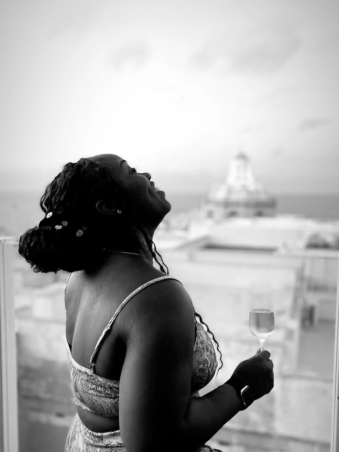 A woman with curly hair holding a wine glass, smiling and looking up, with a blurred building and sky in the background.
