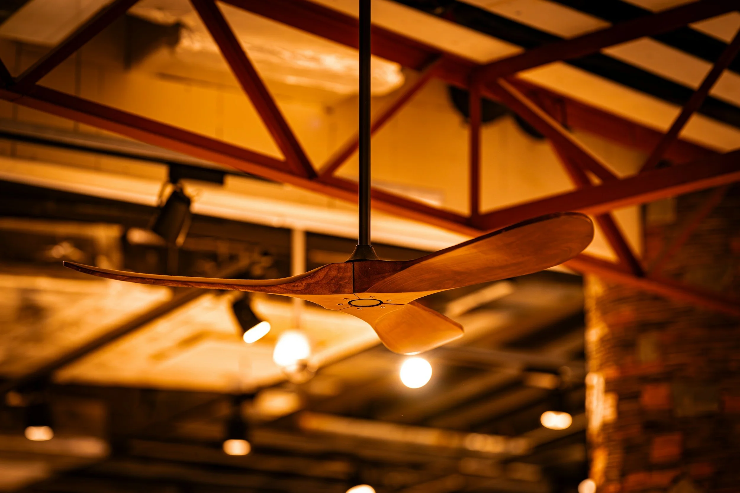 Wooden ceiling fan hanging from ceiling with exposed metal beams and warm lighting.