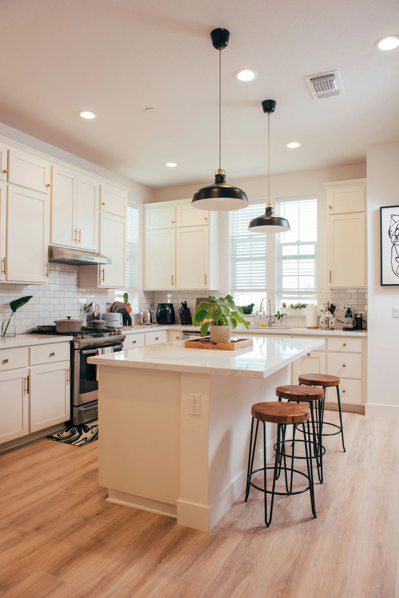Bright kitchen with white cabinets, a marble island with three wooden stools, pendant lights, and a window with blinds.