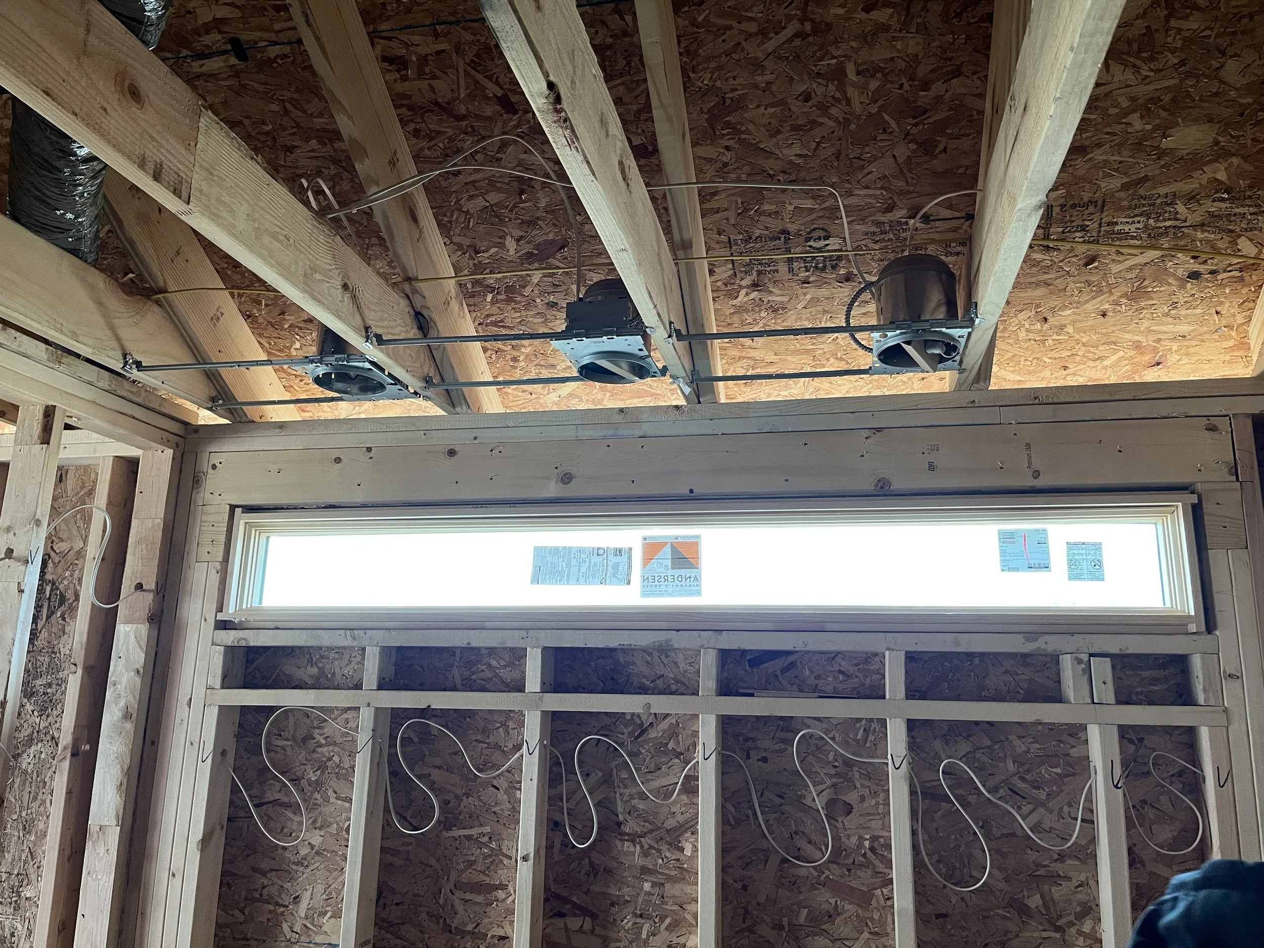 Interior view of a building under construction, showing wooden framing, an OSB ceiling, and a long horizontal window with some stickers. Electrical wiring and ventilation components are visible on the ceiling.