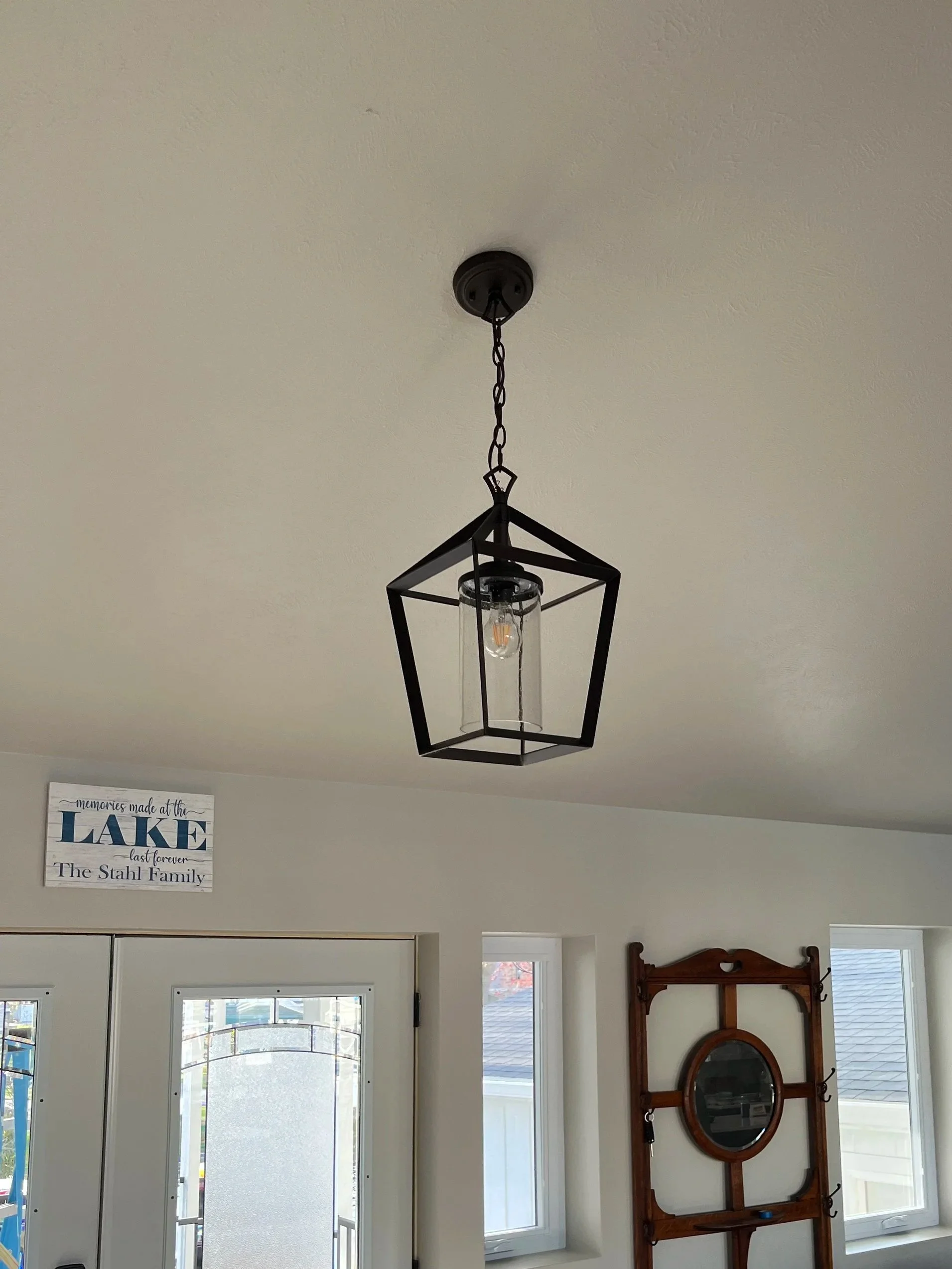A black metal geometric pendant light hanging from the ceiling in a room with white walls, near a white door and windows, and a wooden mirror or coat rack on the wall.
