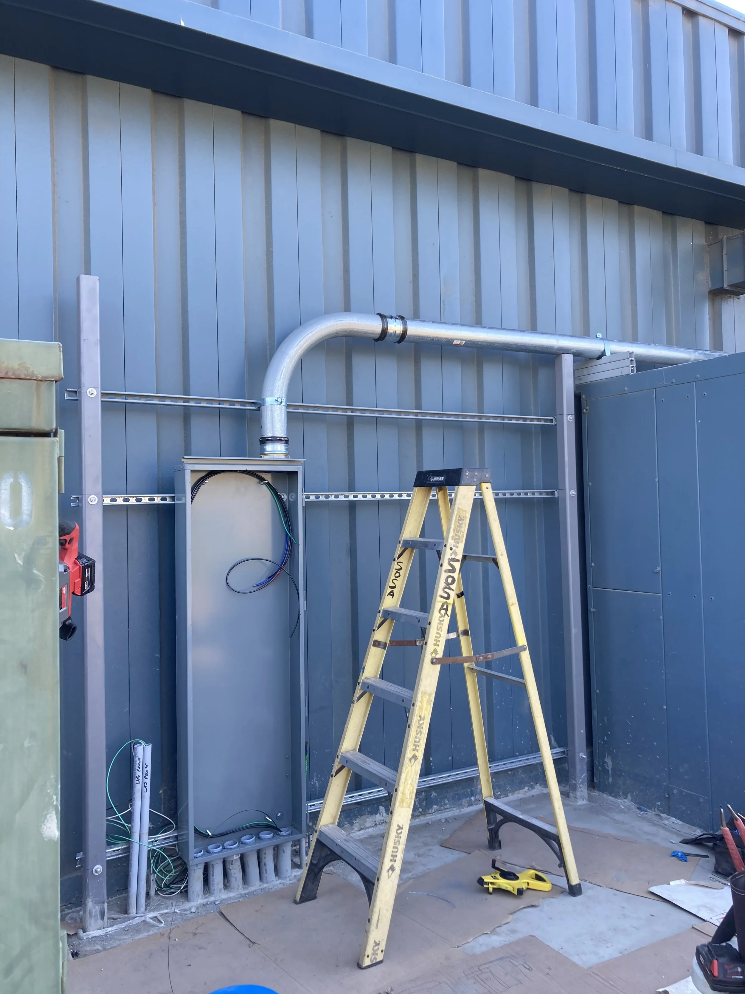Construction site with electrical and ventilation work in progress. A yellow ladder is set up in front of a metallic electrical box and a large pipe attached to the building's blue corrugated metal wall.