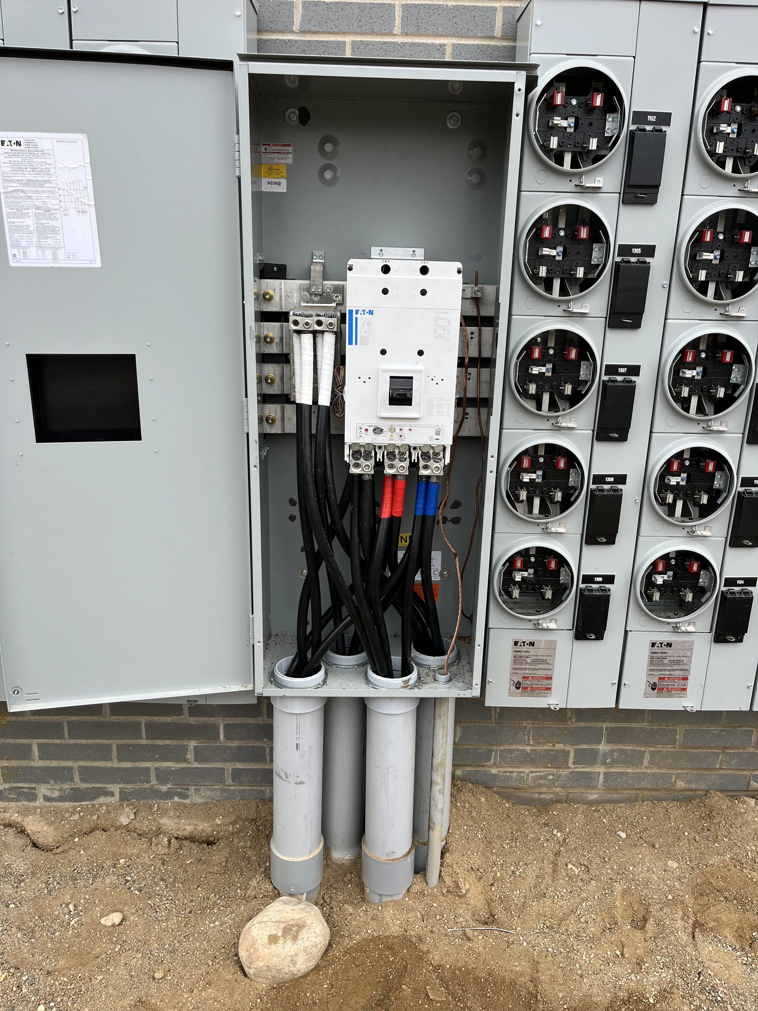 Electrical panel with multiple circuit breakers, wires, and conduits outside on a brick wall. The ground surface is dirt with a large rock in front.