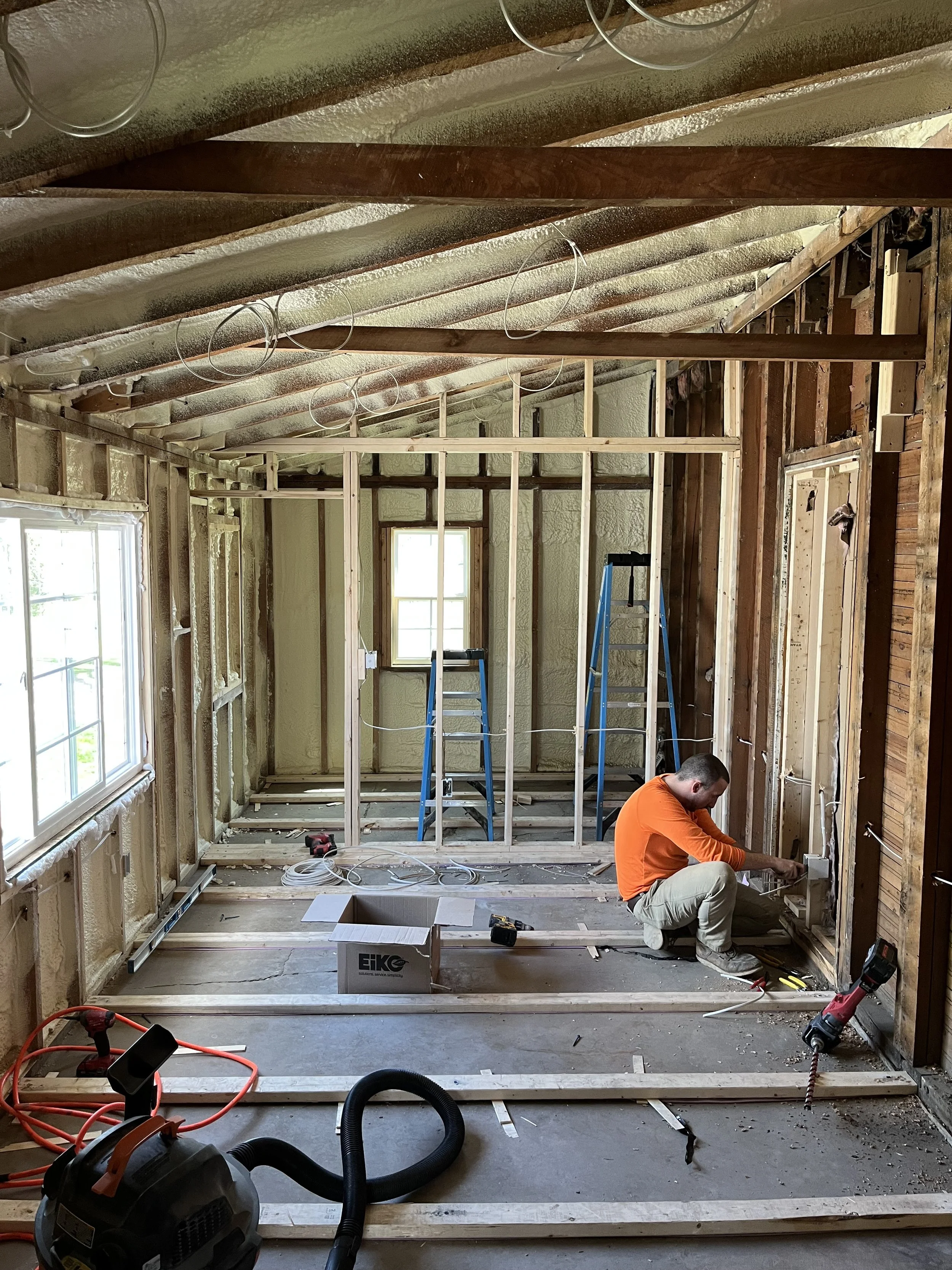 A person working on home renovation, installing electrical wiring and framing in a room with exposed insulation, window, and ceiling beams.