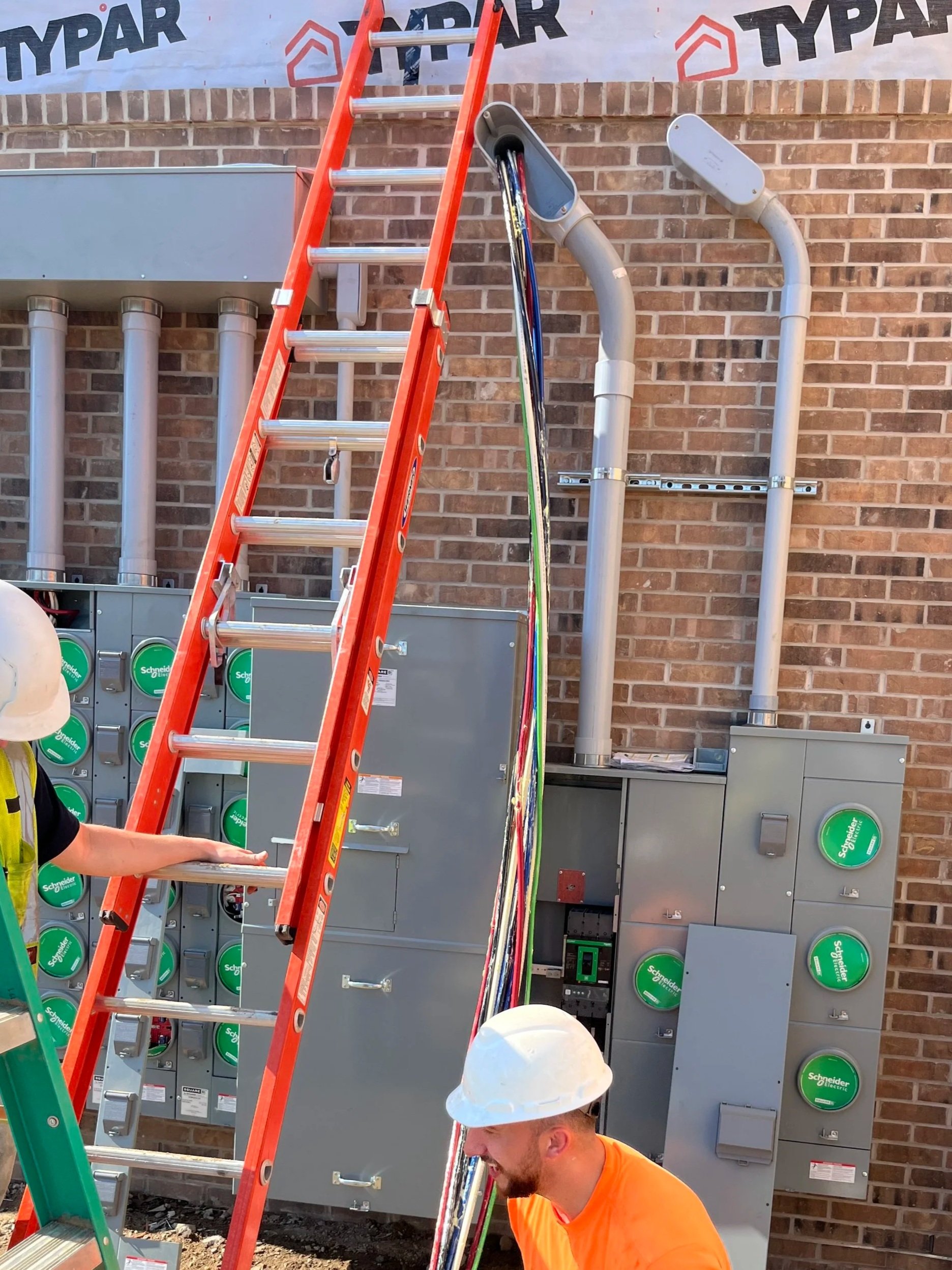 Construction workers installing or maintaining electrical equipment on a brick wall, with a red ladder leaning against a utility panel with green circular labels.
