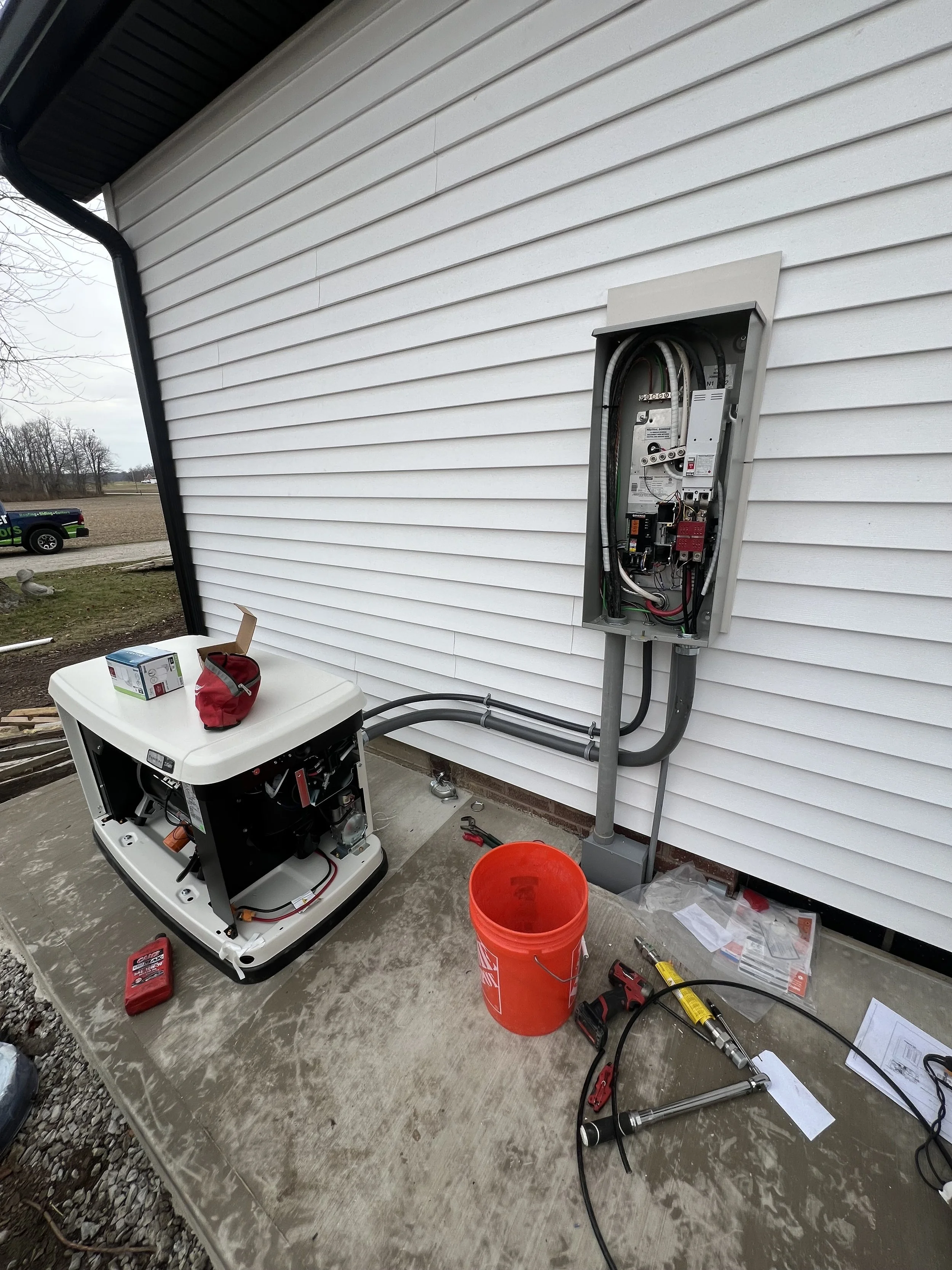 An outdoor electrical panel on a white house wall, with open cover, wires and breakers visible. Near the panel, there's a white generator, tools, a red bucket, and scattered papers on a concrete foundation.