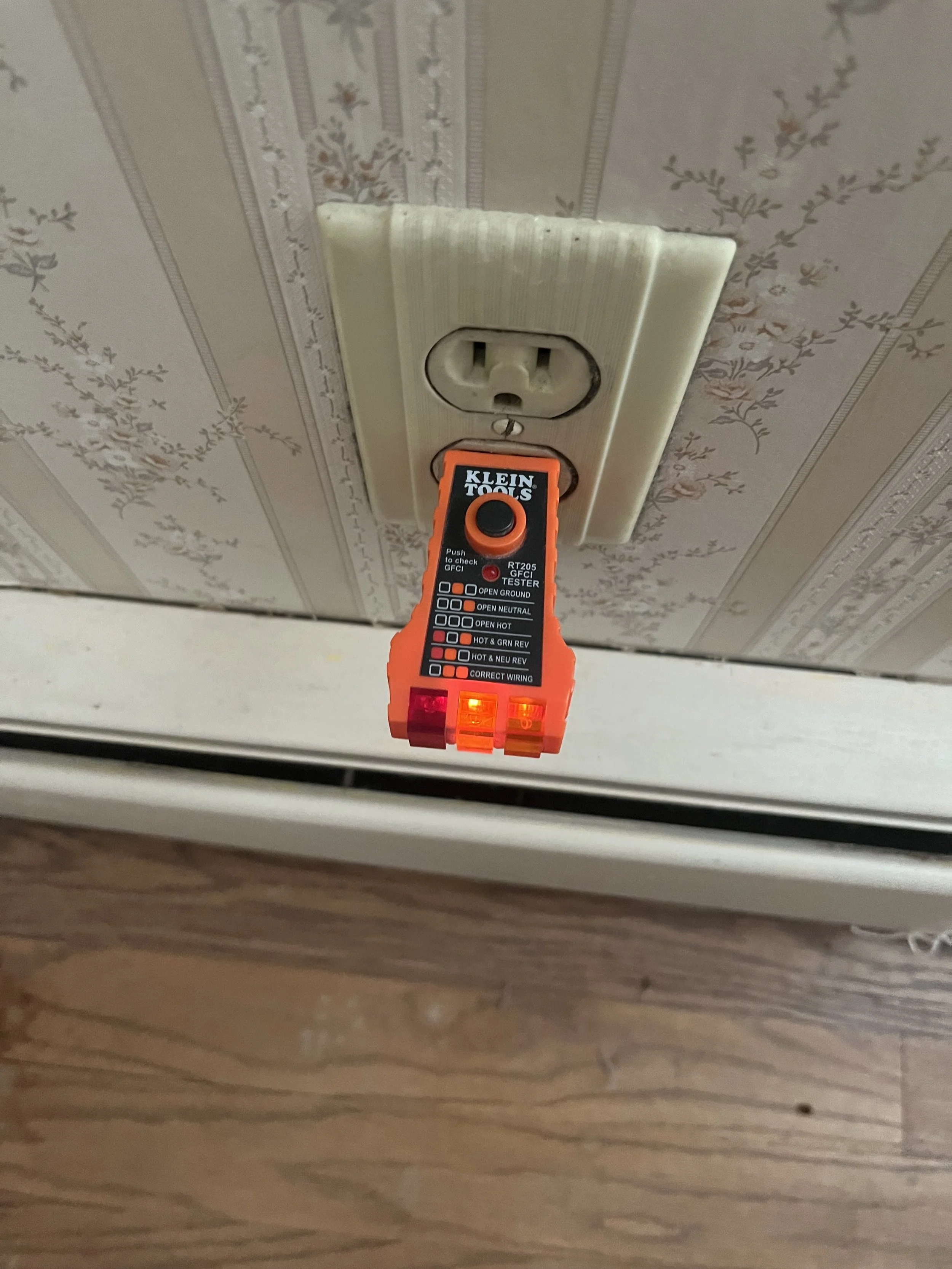 A neon orange circuit Tester hanging from an electrical outlet on a wall with vintage wallpaper, and hardwood flooring below.