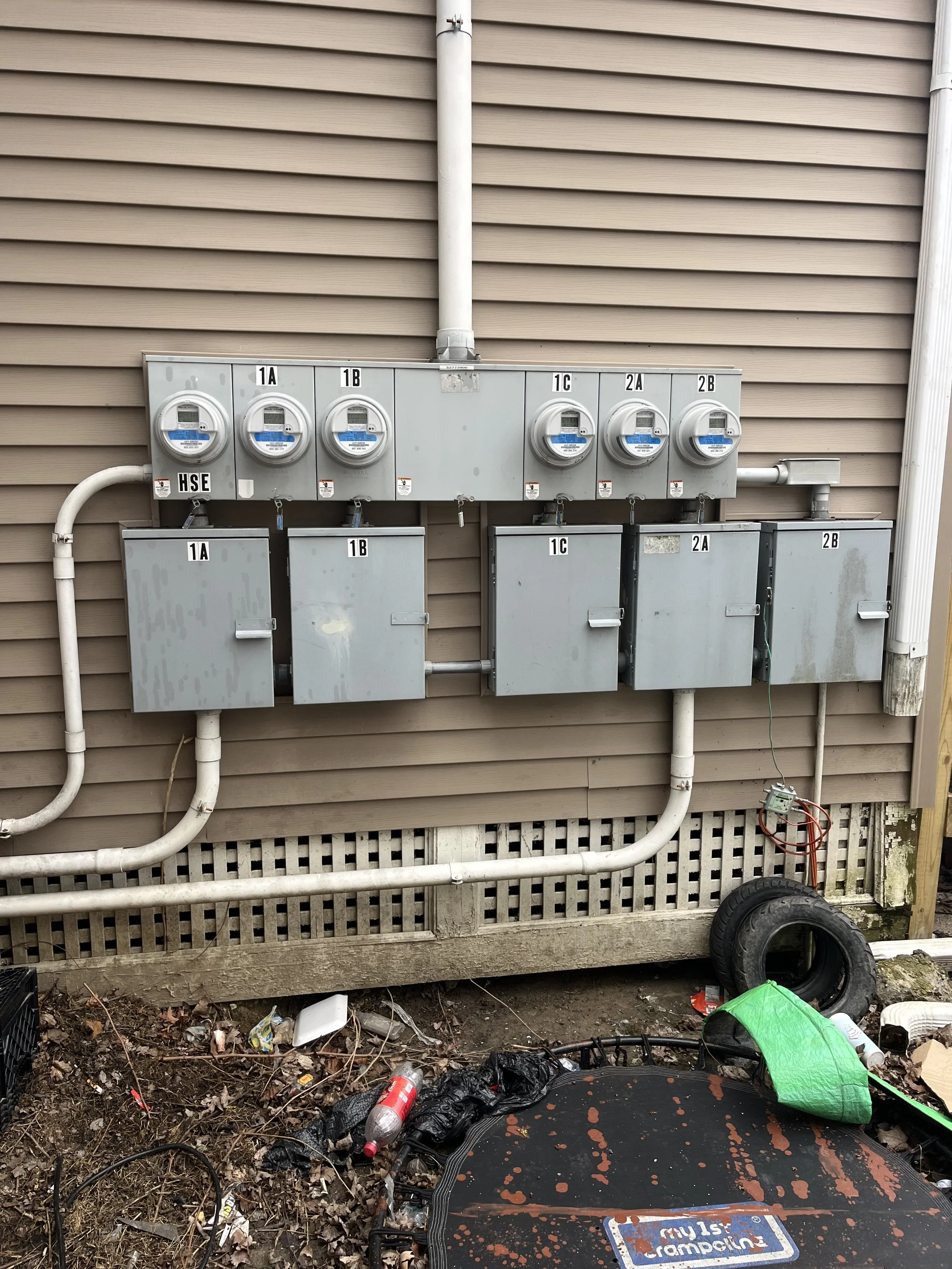 Multiple electrical meters and boxes attached to the beige exterior wall of a building. Some wires and debris scattered on the ground underneath.