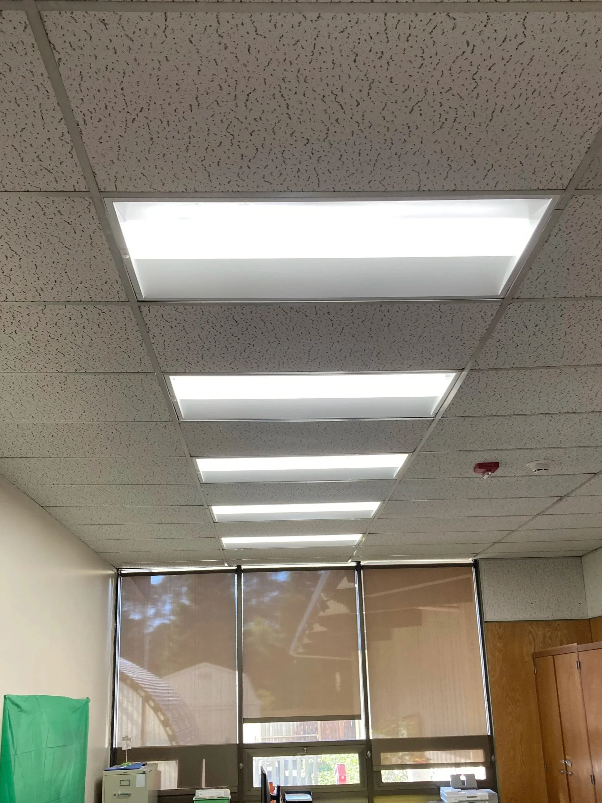 Ceiling with a series of rectangular fluorescent light panels in a room with windows covered by brown blinds, a wooden cabinet, with miscellaneous office supplies.