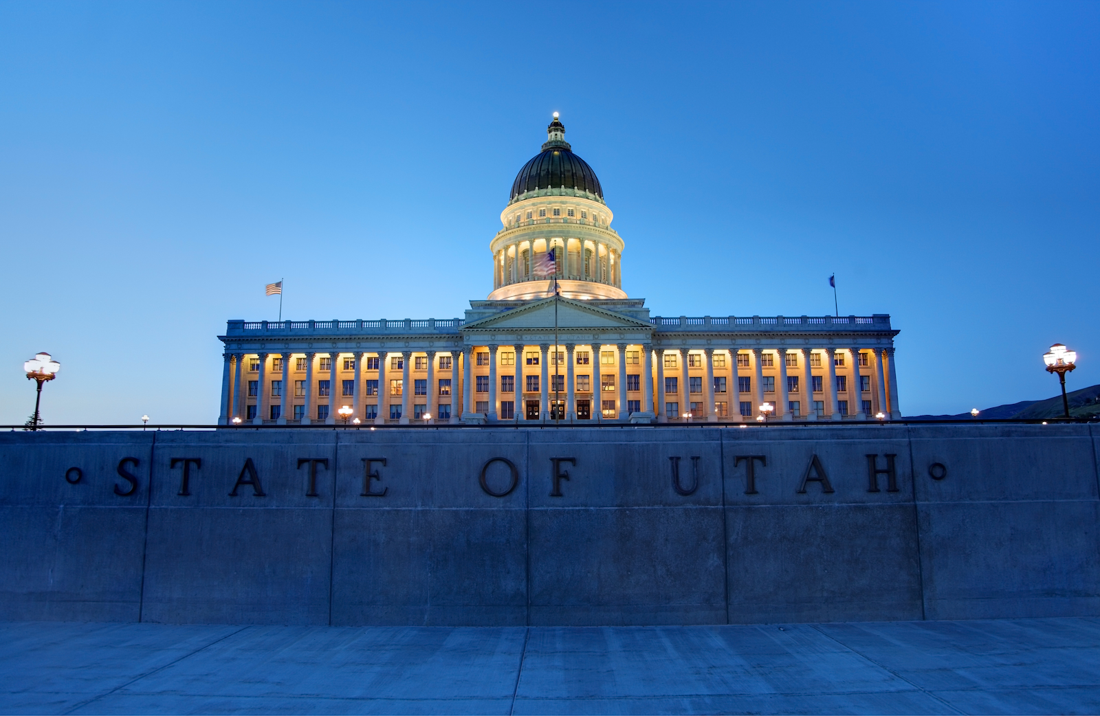 The Utah State Capitol building illuminated at dusk against a clear blue sky, with the words 'STATE OF UTAH' visible on the front wall.