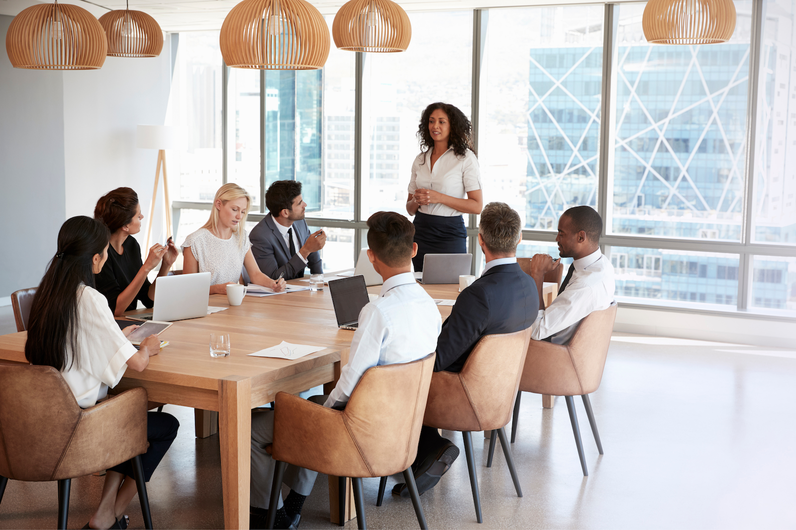 A woman standing and speaking to a group of eight seated professionals in a conference room with large windows and city views.