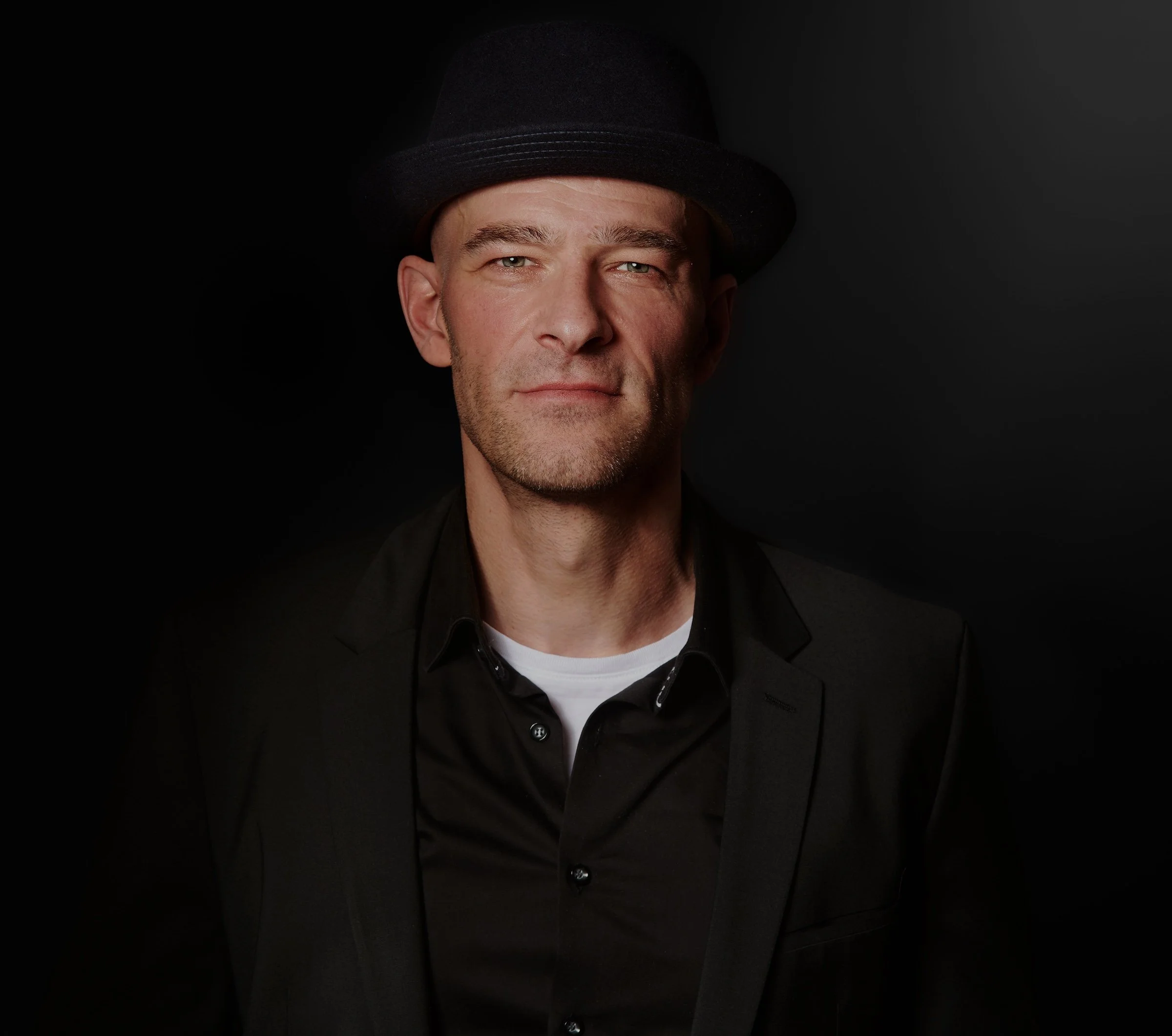 Porträt eines Mannes mit schwarzem Hut, schwarzem Anzug und weißem Hemd vor schwarzem Hintergrund.