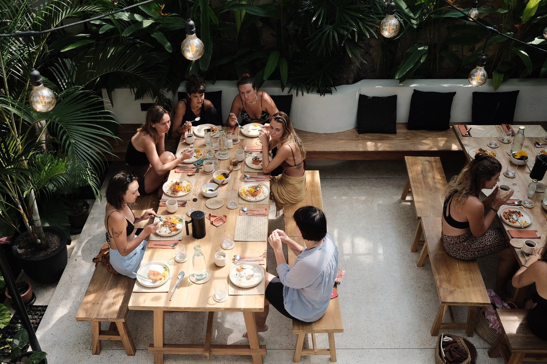 A group of people sitting at wooden tables in a restaurant with lush green plants, enjoying a meal.