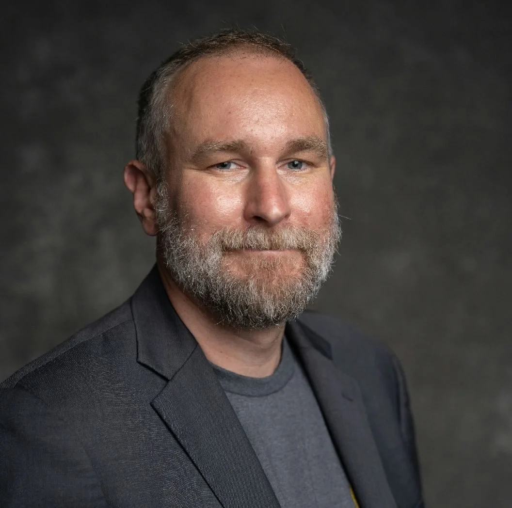 A professional woman with long dark hair, wearing a rust-colored blazer and holding a disposable coffee cup and a brown notebook, standing indoors near large windows with greenery outside.