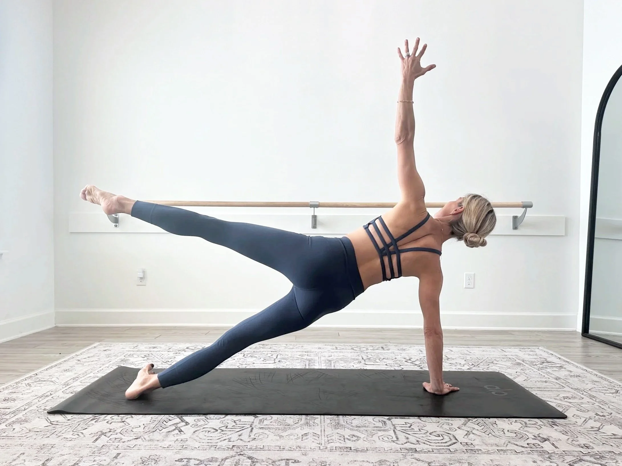 A woman doing a side plank yoga pose in a dance studio with ballet barres and a mirror.