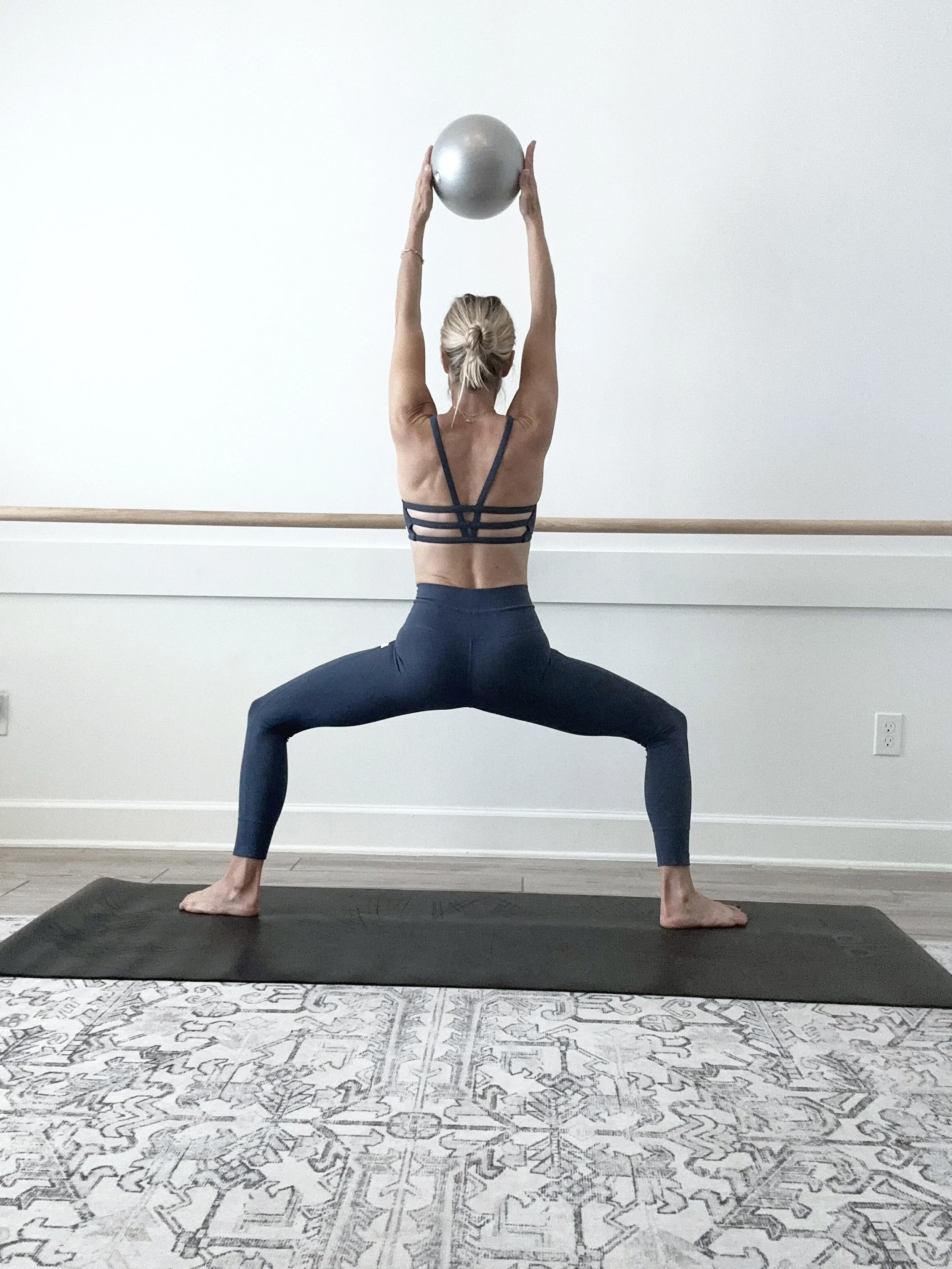 A woman practicing yoga in a lunge pose with wide legs and arms raised overhead holding a silver exercise ball, on a black yoga mat in a minimalistic room.
