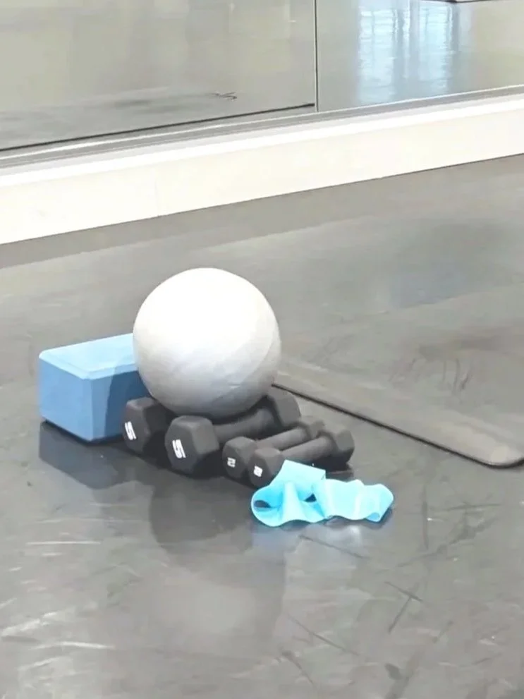 A fitness setup on the floor including a white exercise ball, two blue workout blocks, three small black dumbbells, a blue resistance band, and a gray exercise mat.