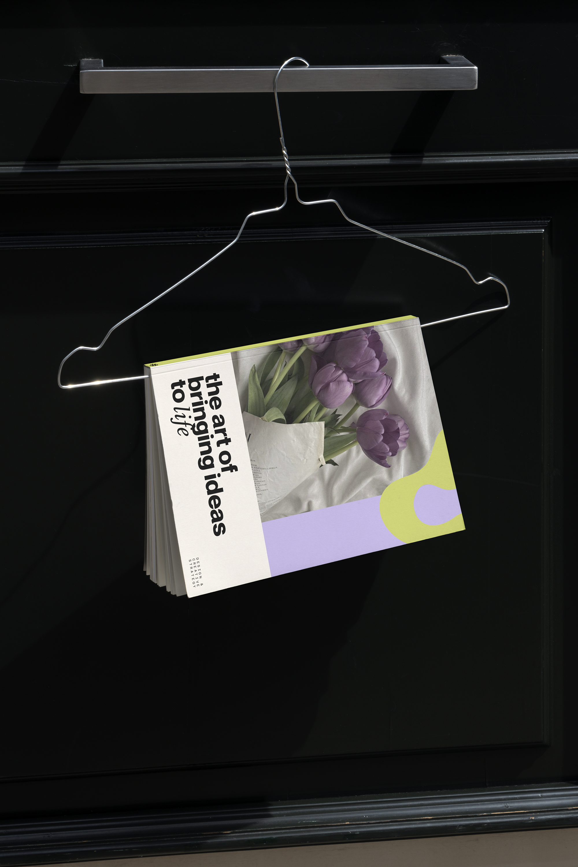 A magazine with purple tulips on the cover and black text, resting on a black surface beside a metal bar and a glass surface.