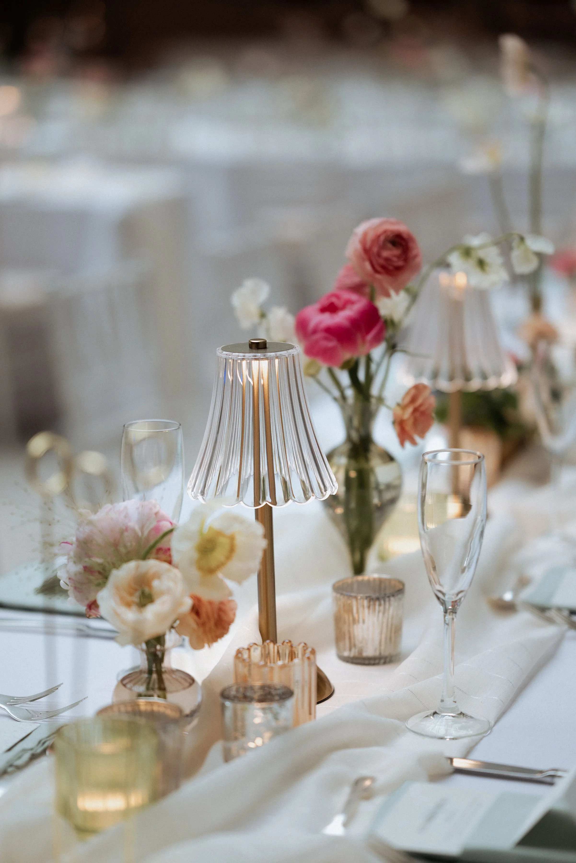 Elegant table setting with pink and white flowers, candle holders, and a small lamp on a white tablecloth.