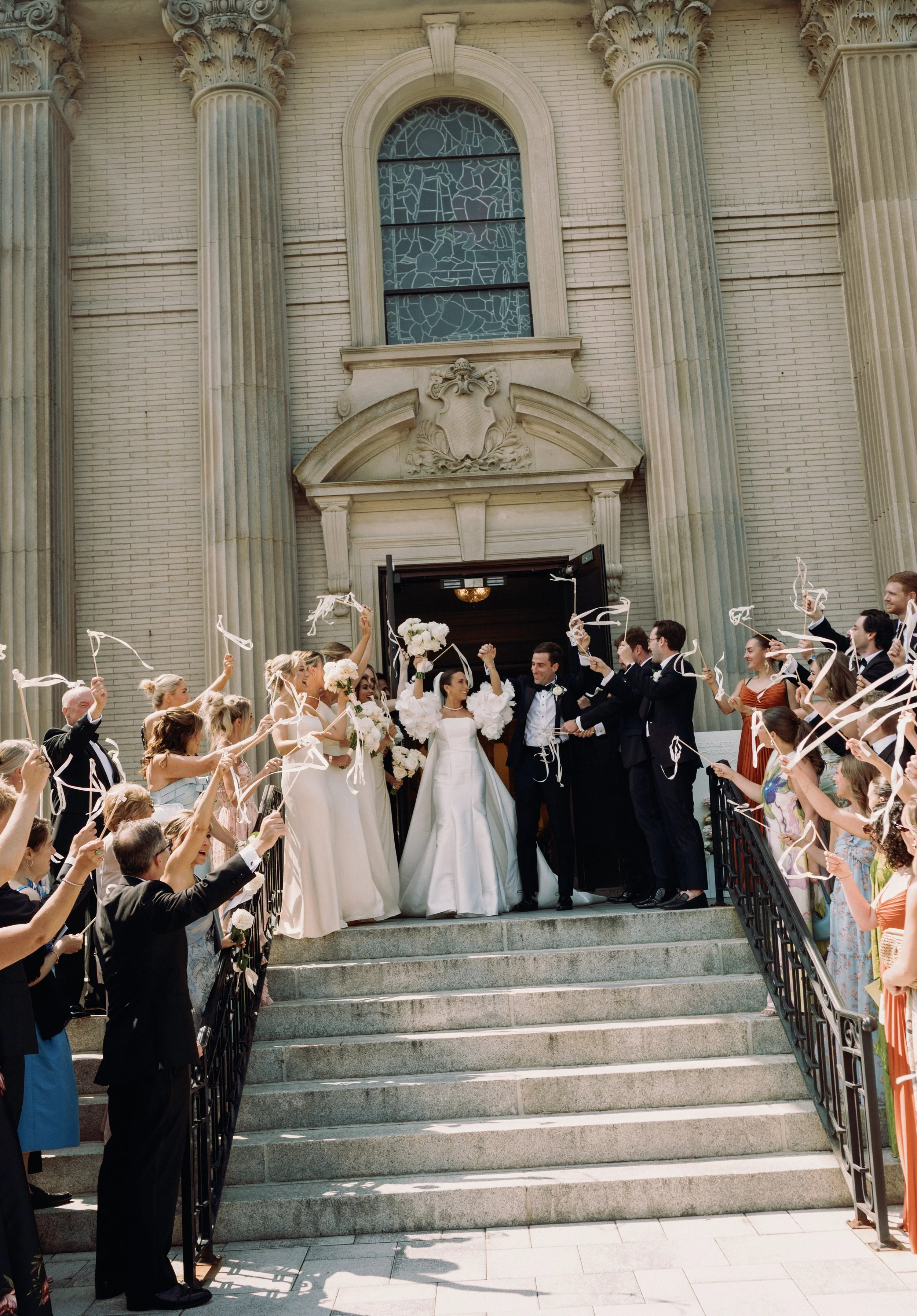 Wedding celebration outside a church with the bride and groom on top of stairs, surrounded by friends and family waving ribbons and celebrating.