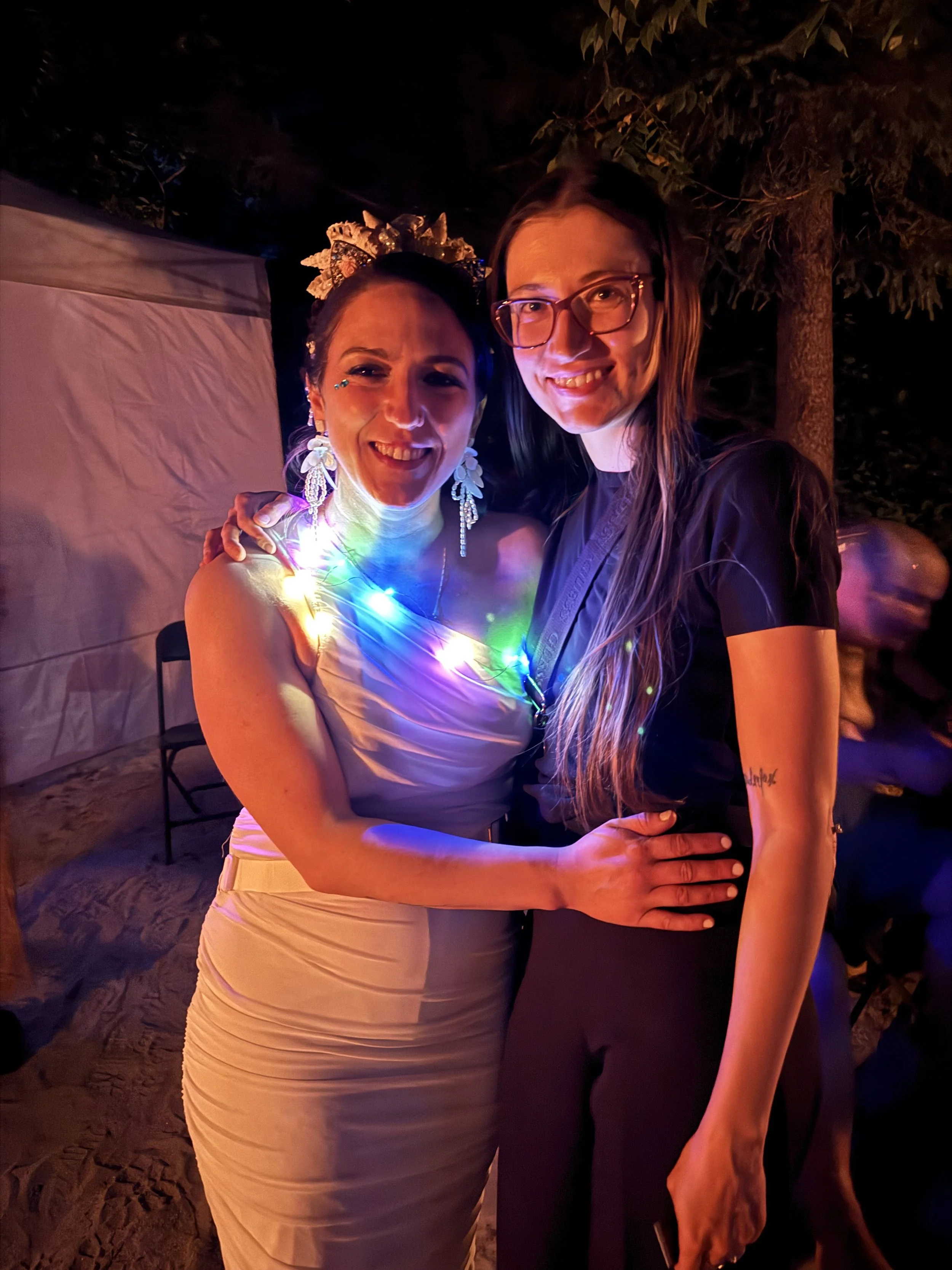 Two women smiling and embracing at an outdoor night event, one wearing a white dress with colorful LED lights around her neck, and the other wearing glasses and a dark outfit, with trees and blurred figures in the background.