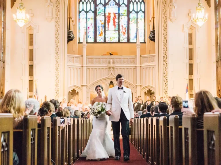 Bride and groom walking down the aisle inside a church during their wedding ceremony, with guests seated on both sides and stained glass windows in the background.