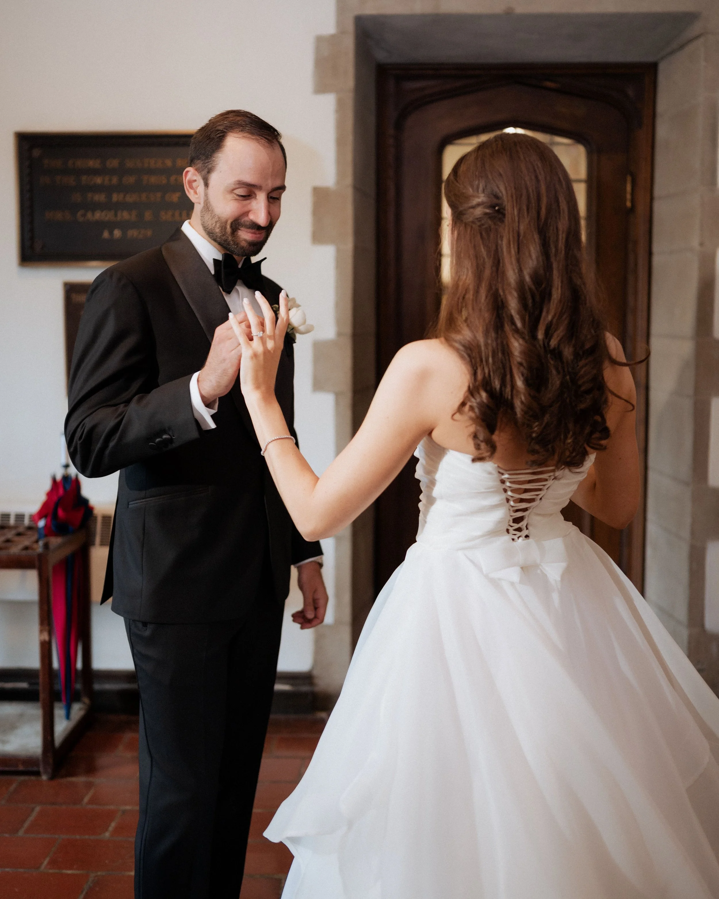 A bride and groom are dancing at their wedding, holding hands and smiling. The bride is wearing a white wedding gown with a lace-up back, and the groom is dressed in a black tuxedo with a bow tie. They are indoors, near a wooden door and a plaque on the wall.